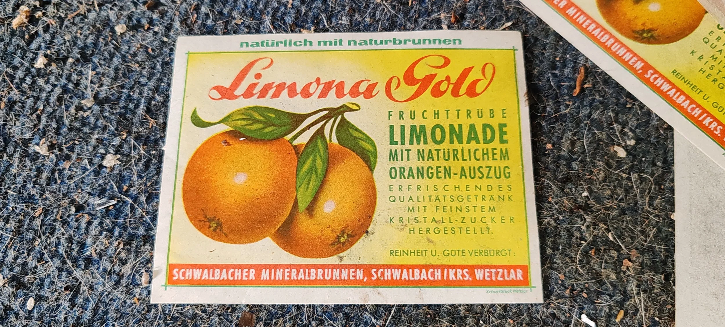 Etikett für Limona Gold Fruchtrübe Limonade mit Orangenauszug auf einer dunkel gemusterten Oberfläche, mit Illustration von Orangen und grünen Blättern.