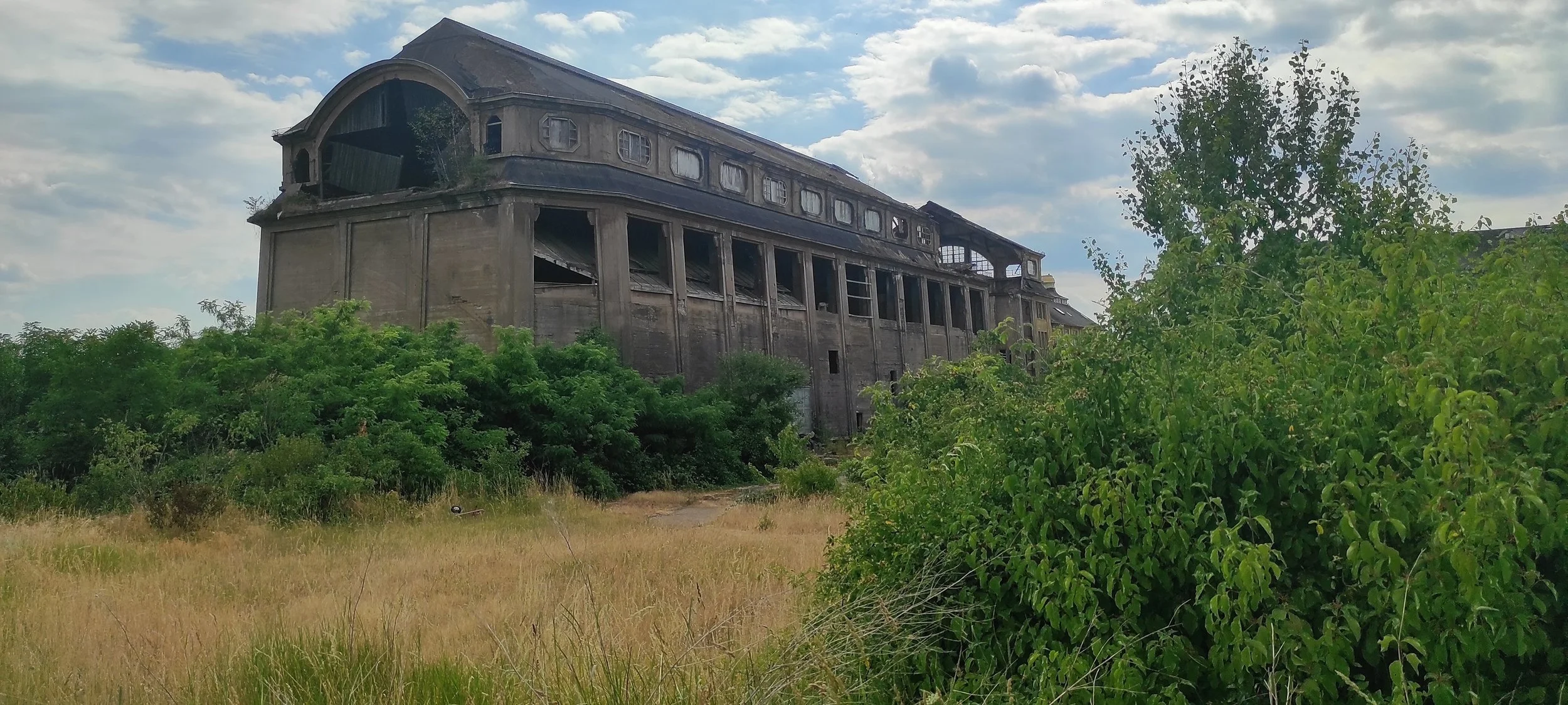 Verfallenes, verlassenes und verfallenes Gebäude auf einer Wiese mit Büschen und Bäumen im Vordergrund, Himmel mit Wolken im Hintergrund.