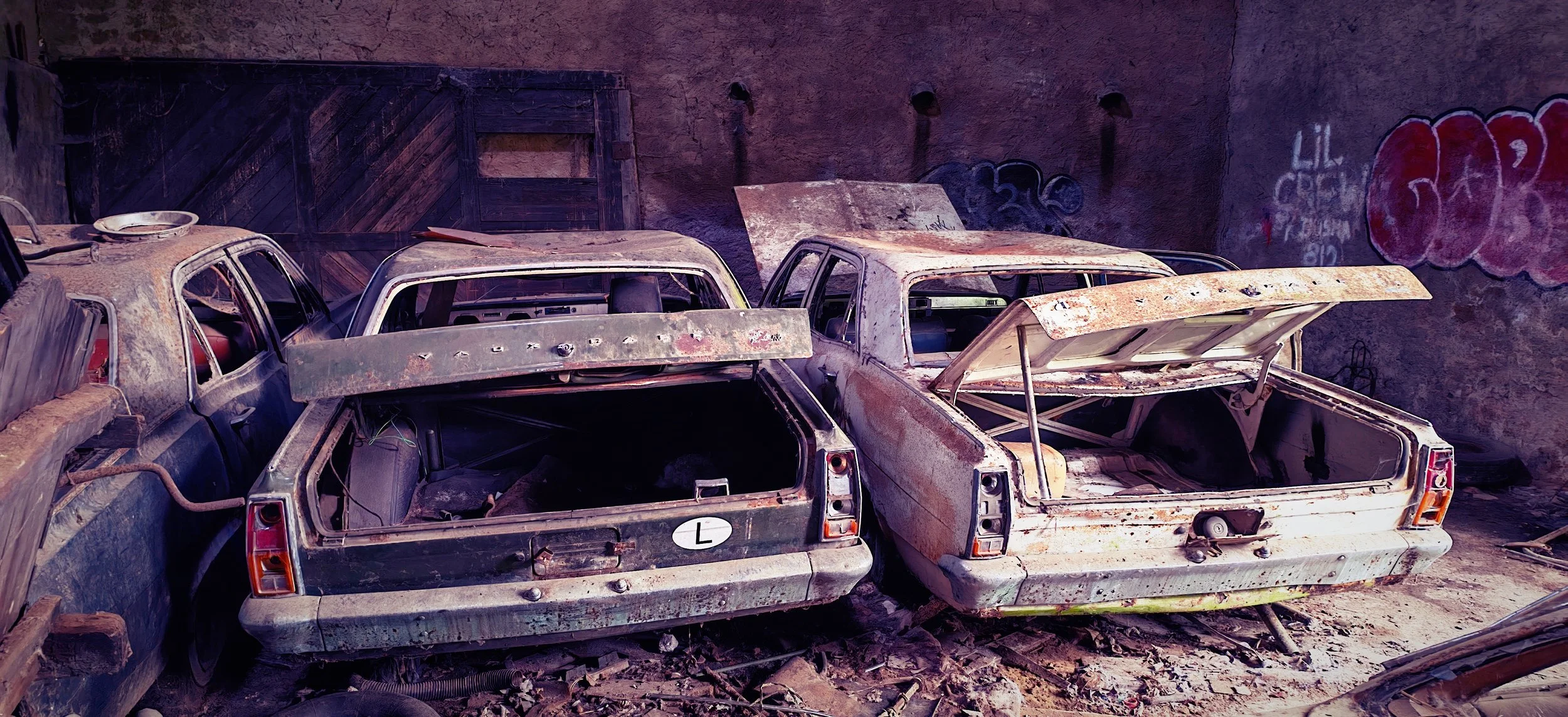 Ein Raum mit mehreren alten, verrosteten Autos, die ineinander gestapelt sind, vor einer Wand mit Graffiti.