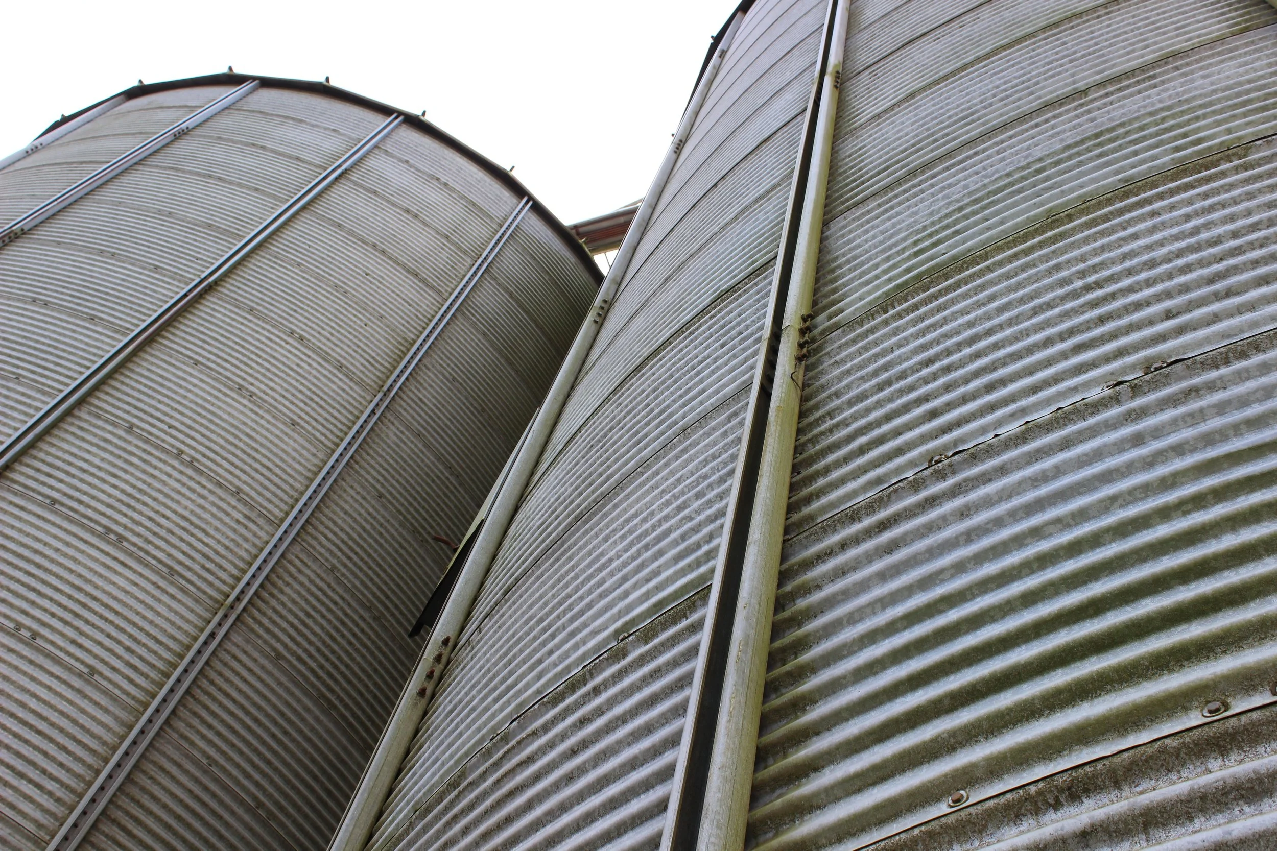Blick auf die Seiten von zwei großen Getreidesilos aus Metall, aufgenommen von unten, gegen den Himmel.