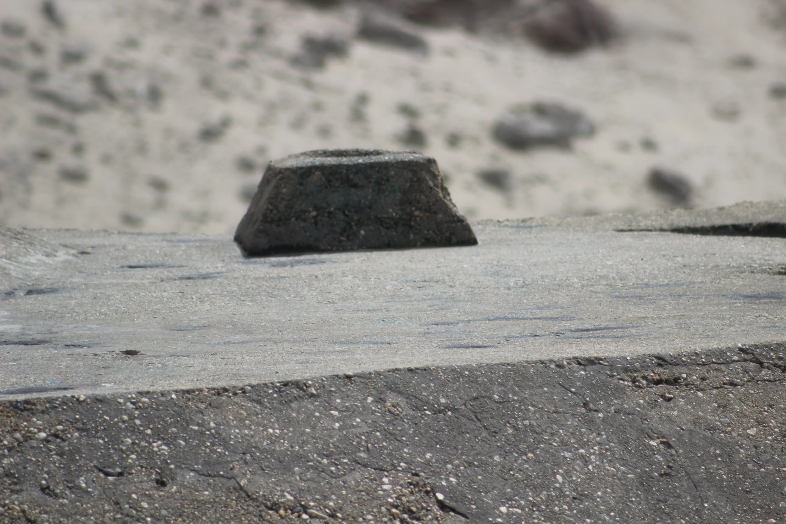 Ein asphaltierter Bürgersteig mit einer abstehenden Kante, im Hintergrund Sand und kleine Steine