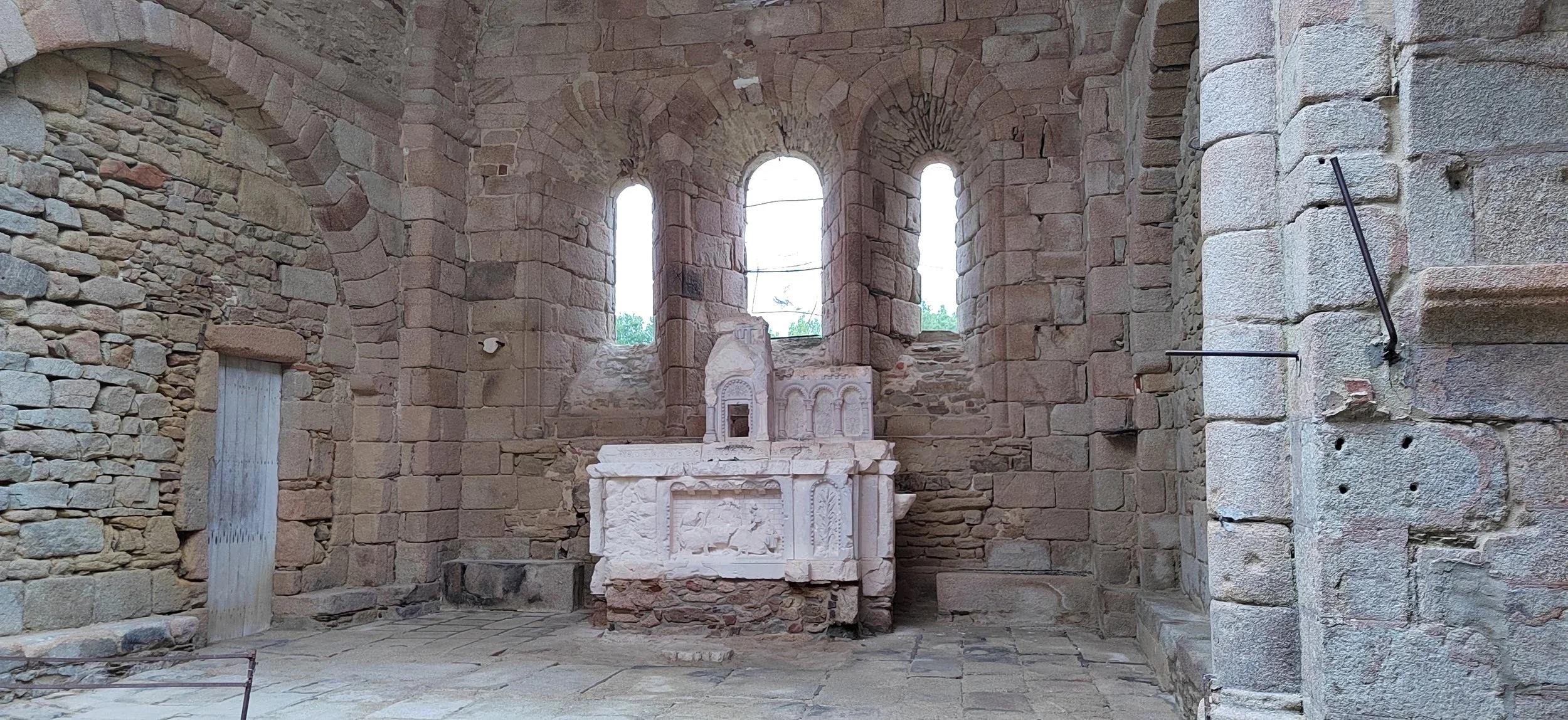 Innenseite einer alten Kirche mit Steinmauer, drei runde Fenster und einem alten Altar aus Stein.
