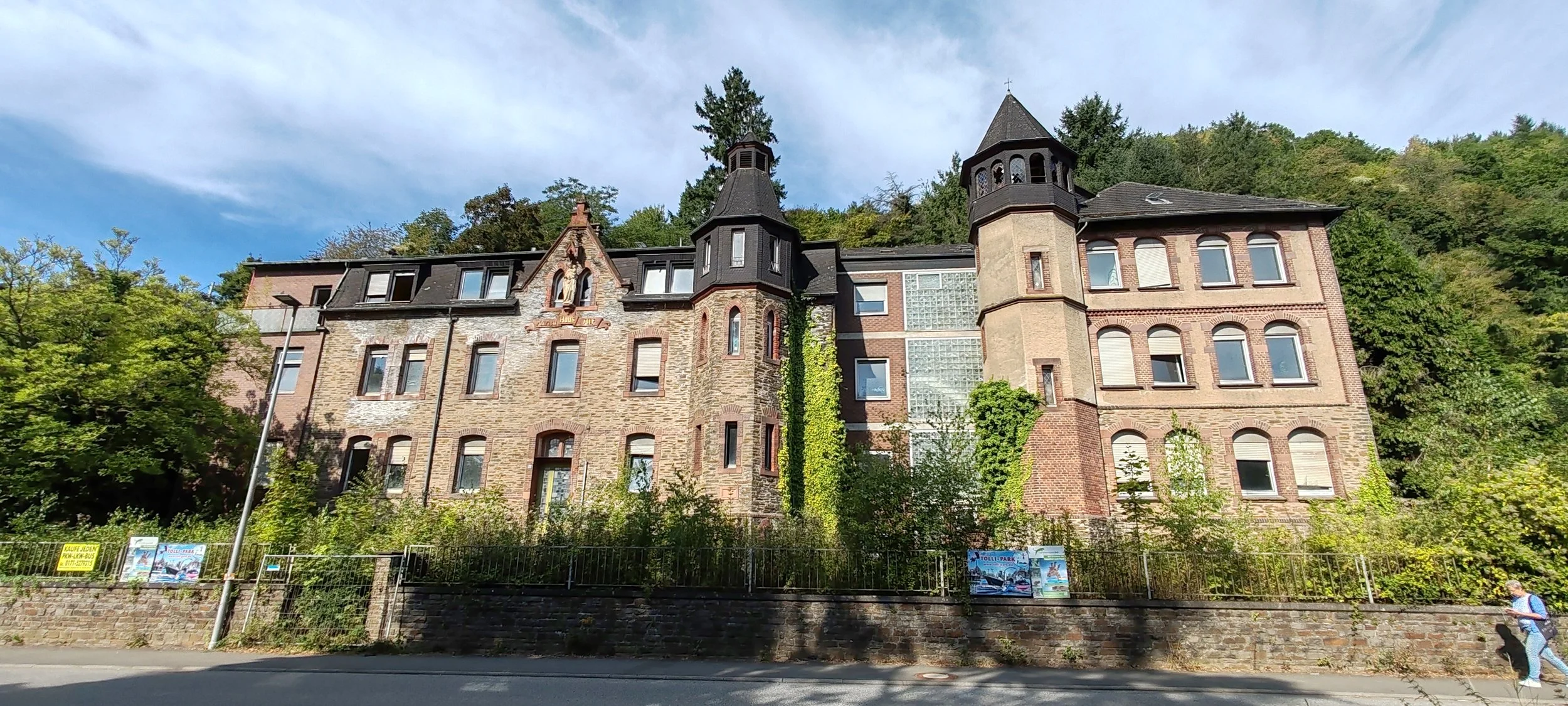 Historisches Gebäude mit Türmen und vielen Fenstern, umgeben von Bäumen, im Hintergrund ein bewölkter Himmel.