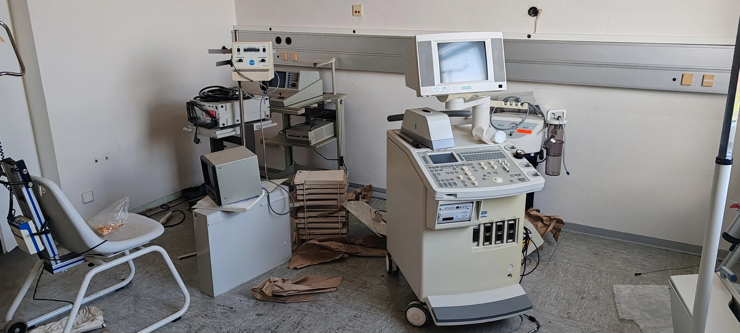 Un veraltetes, chaotisches medizinisches Untersuchungszimmer mit einem alten Ultraschallgerät, einem Stuhl, Papier und technischen Geräten, alles sieht unordentlich aus.