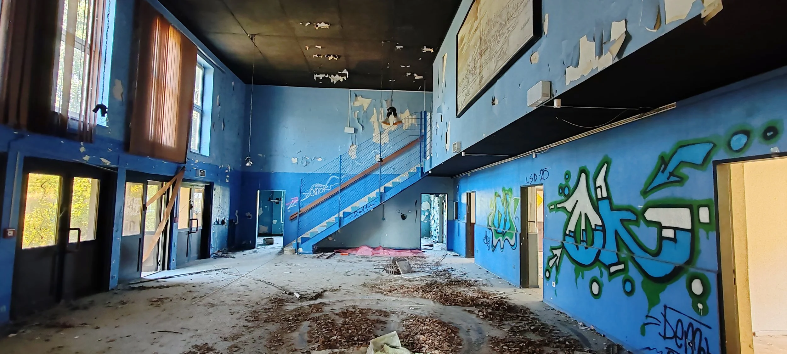 Innenansicht eines verlassenen, verwahrlosten Gebäudes mit blauer Wandfarbe, Graffiti an der Wand, zerbrochenen Fenstern, eine Treppe mit blauer Geländer, beschädigte Decke und aufgerissenem Boden.