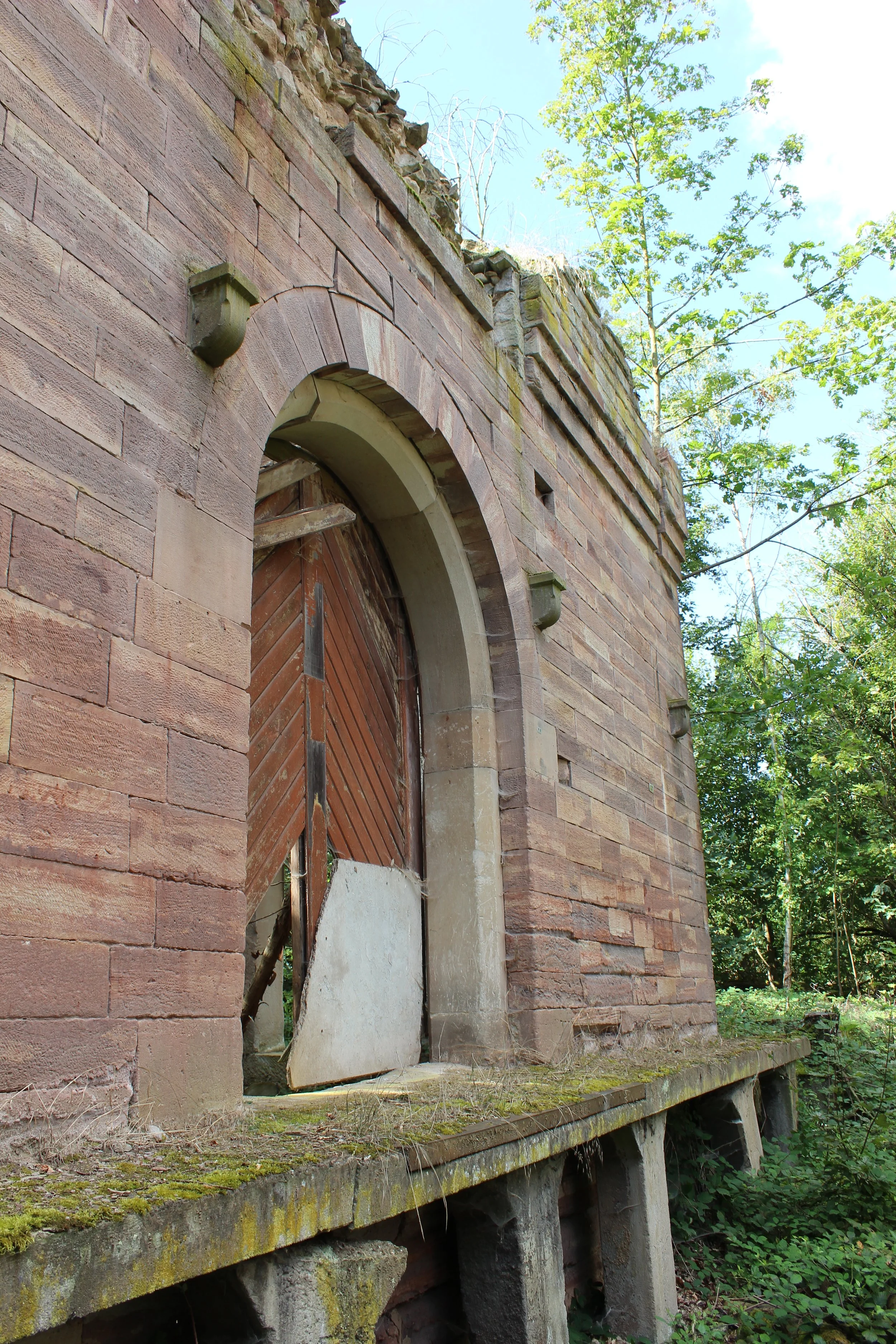Alte, verlassene Feldtor in einem verfallenen, auf einem Betonsockel stehenden Gebäude aus rotem Sandstein, umgeben von Wald und Bäumen.