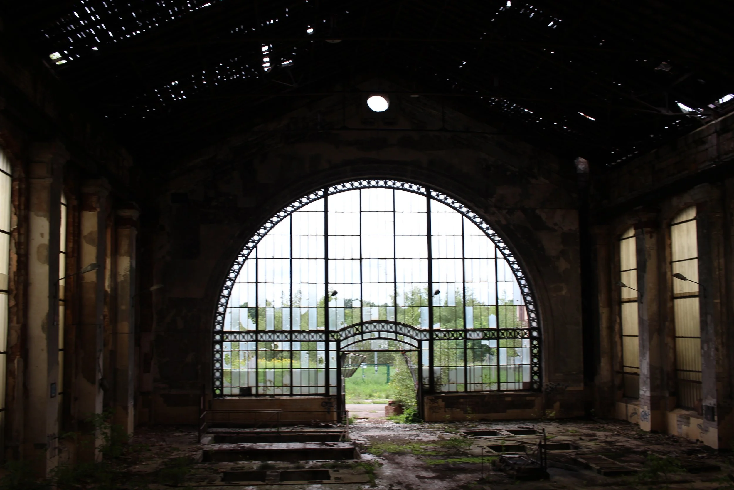 Verlassene, verfallene alte Fabrikhalle mit großem, halb geöffnetem Metalltor und großen Fenstern, durch die man das grüne Freigelände sieht