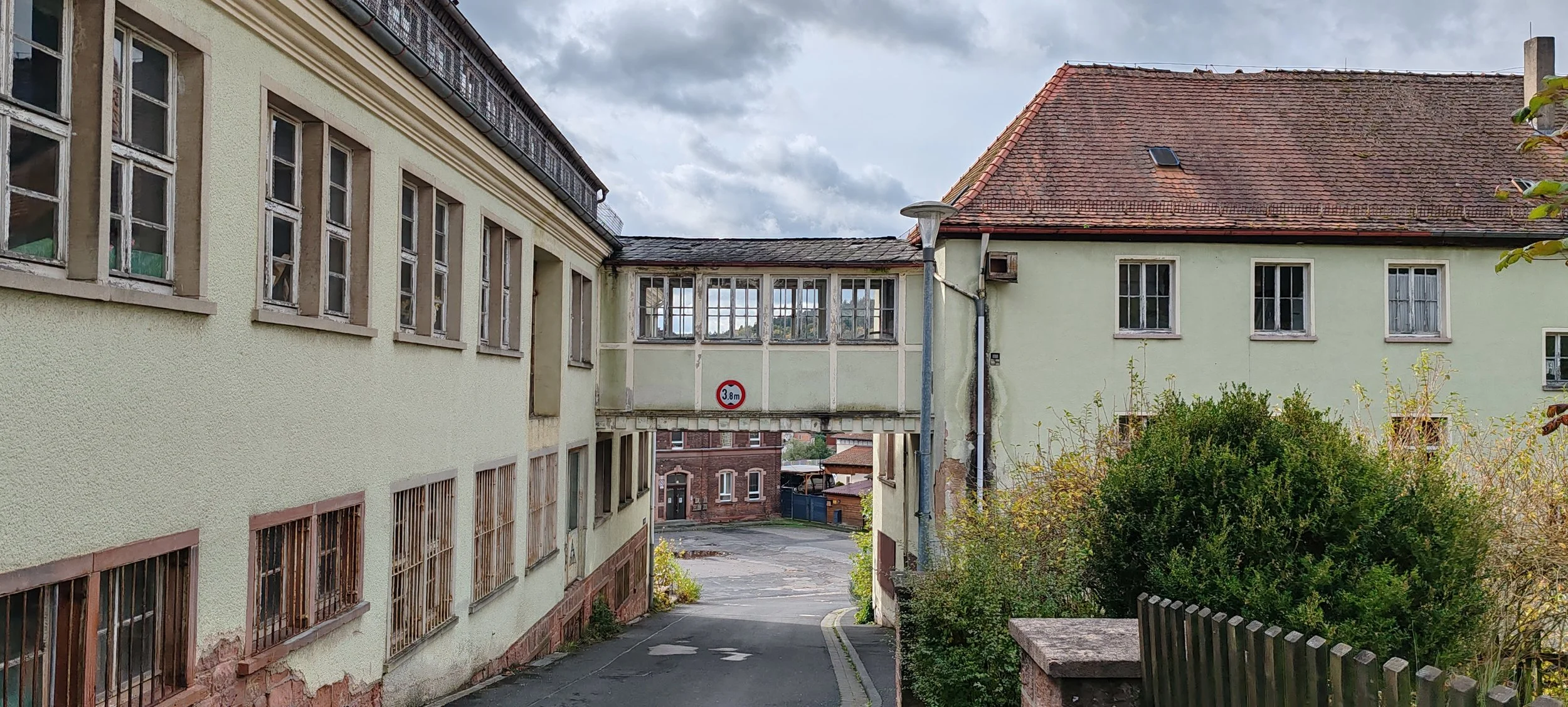 Ein Blick auf ein Gebäude mit grün gestrichener Fassade, einem Durchgang mit einer Höhenbegrenzung von 3,8 Metern, und einem Haus mit rotem Ziegeldach. Im Vordergrund sind Büsche und eine kleine Mauer zu sehen.