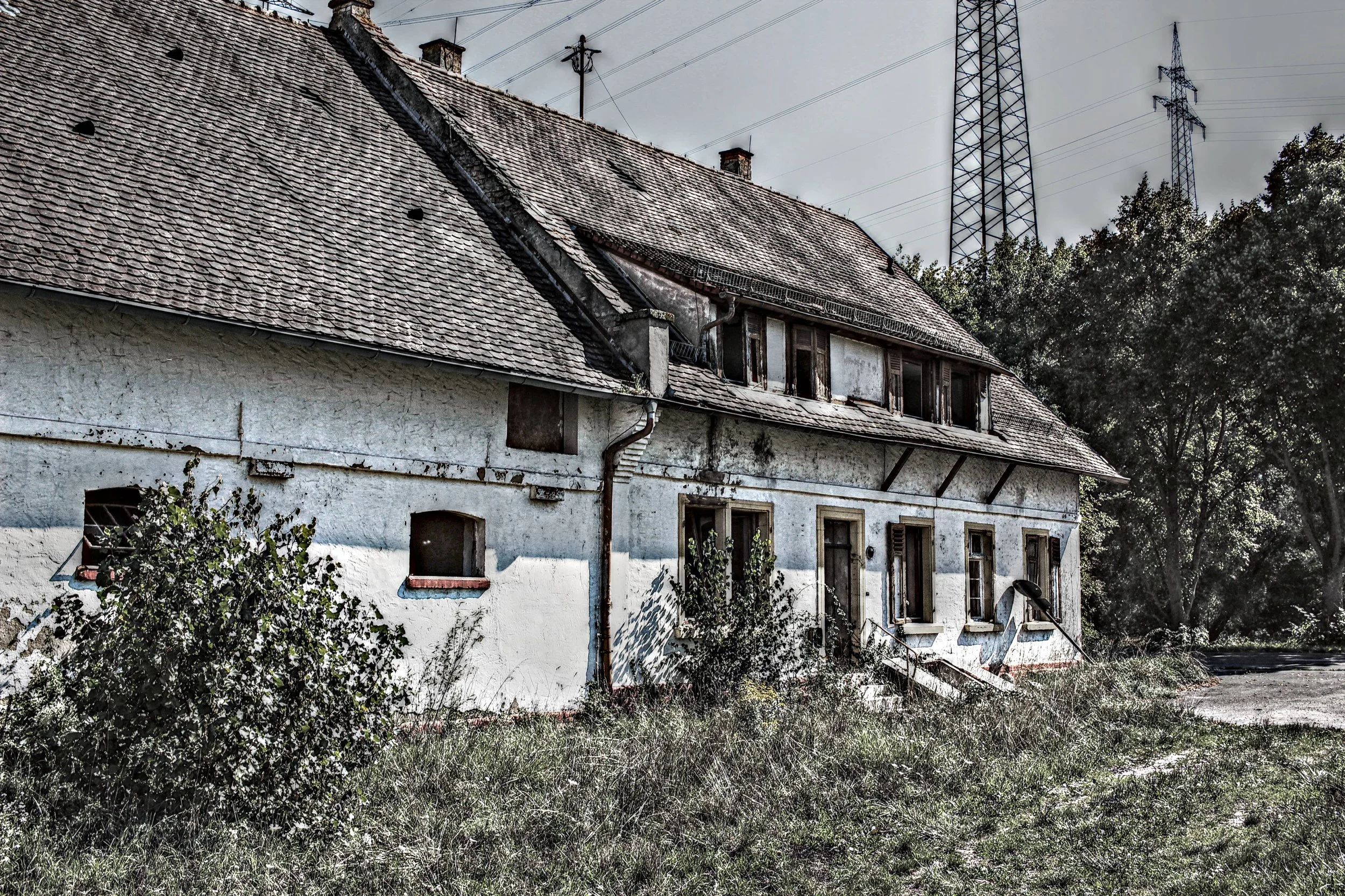 Ein verfallenes, altes Haus mit beschädigtem Dach und verwildertem Garten, im Hintergrund sind Hochspannungsmasten zu sehen.