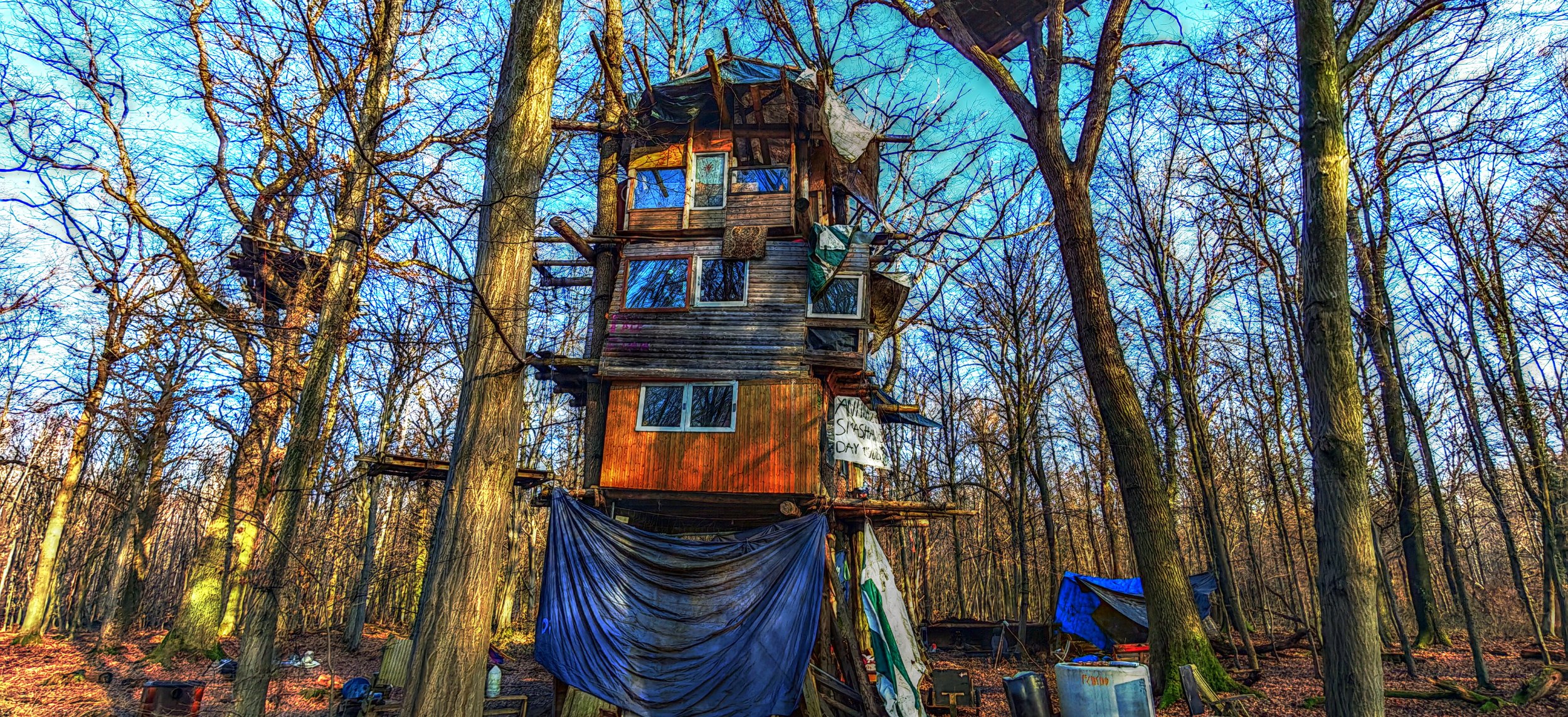 Ein selbstgebautes Baumhaus in einem Winterwald, umgeben von Bäumen ohne Blätter, mit verschiedenen Materialien und Gemälden daran, darunter ein Schild mit der Aufschrift 'Never Surrender Day'