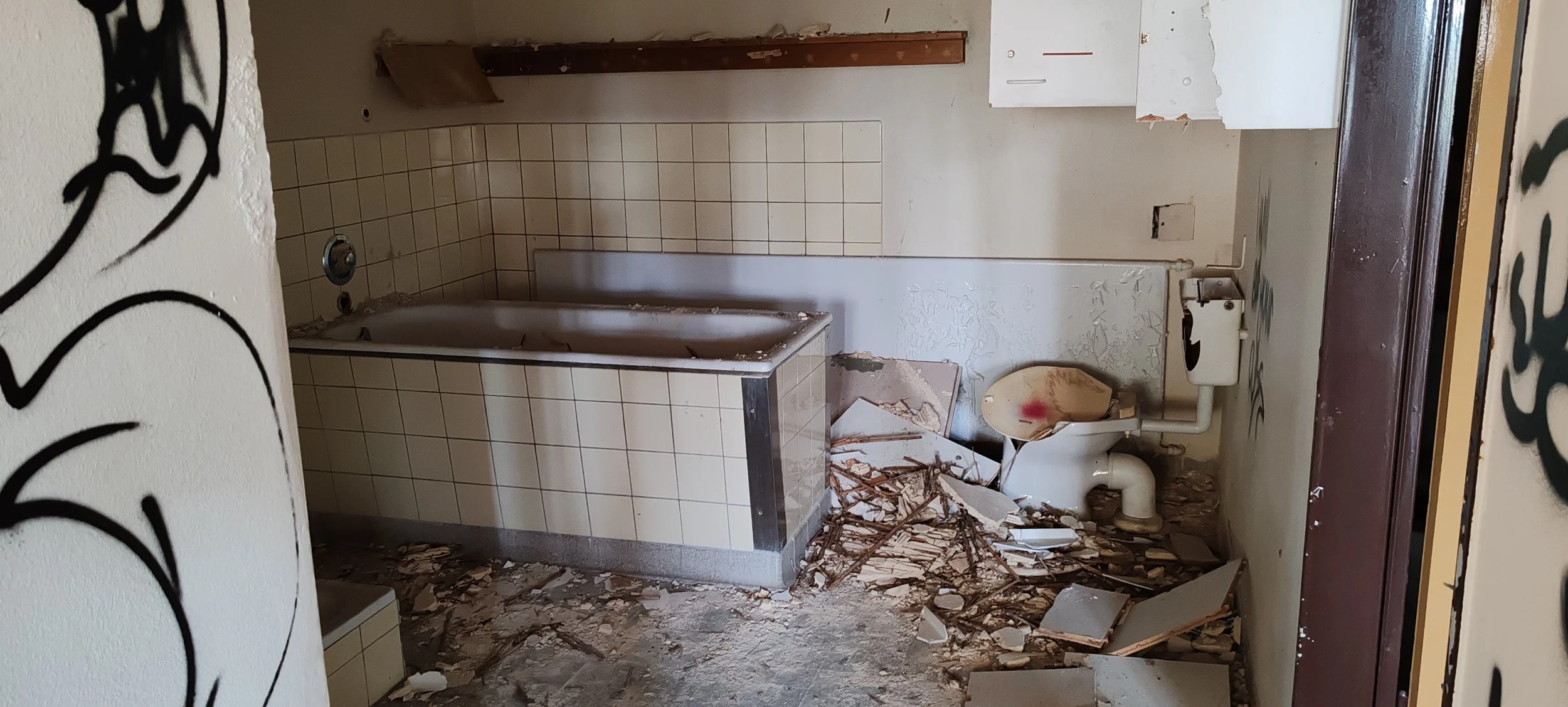 Ein verfallenes Badezimmer mit zerstörtem WC, umgeben von Trümmern und Schmutz, wobei eine Wanne an der Wand steht.