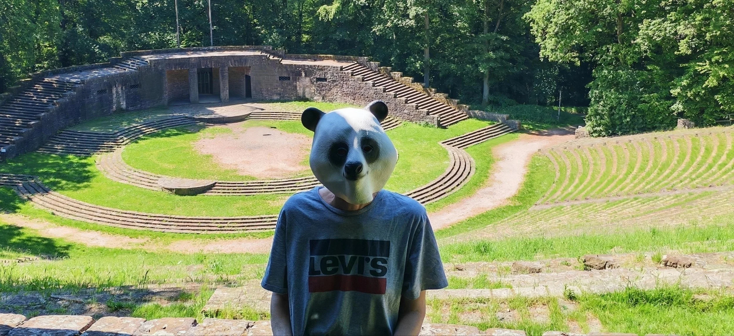 Kind mit Panda-Maske vor antiker Amphitheater im Grünen.