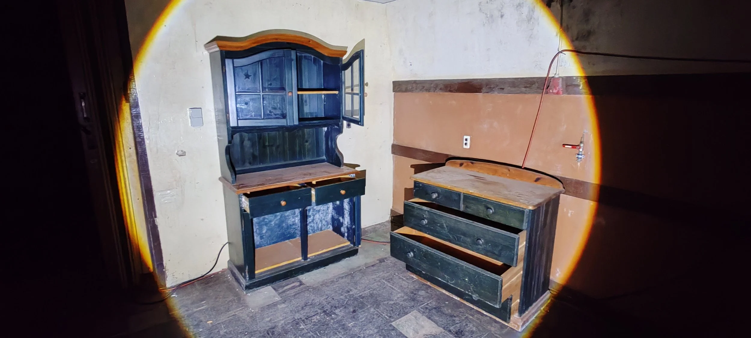 Abgeblendete Ecke eines Raums mit alten, dunklen Möbeln, eine Kommode und ein Regal, auf einer Wand leuchtet eine leere Wandsteckdose, auf dem Boden liegen einige Steine, eine Wasserleitung ist sichtbar an der Wand. 