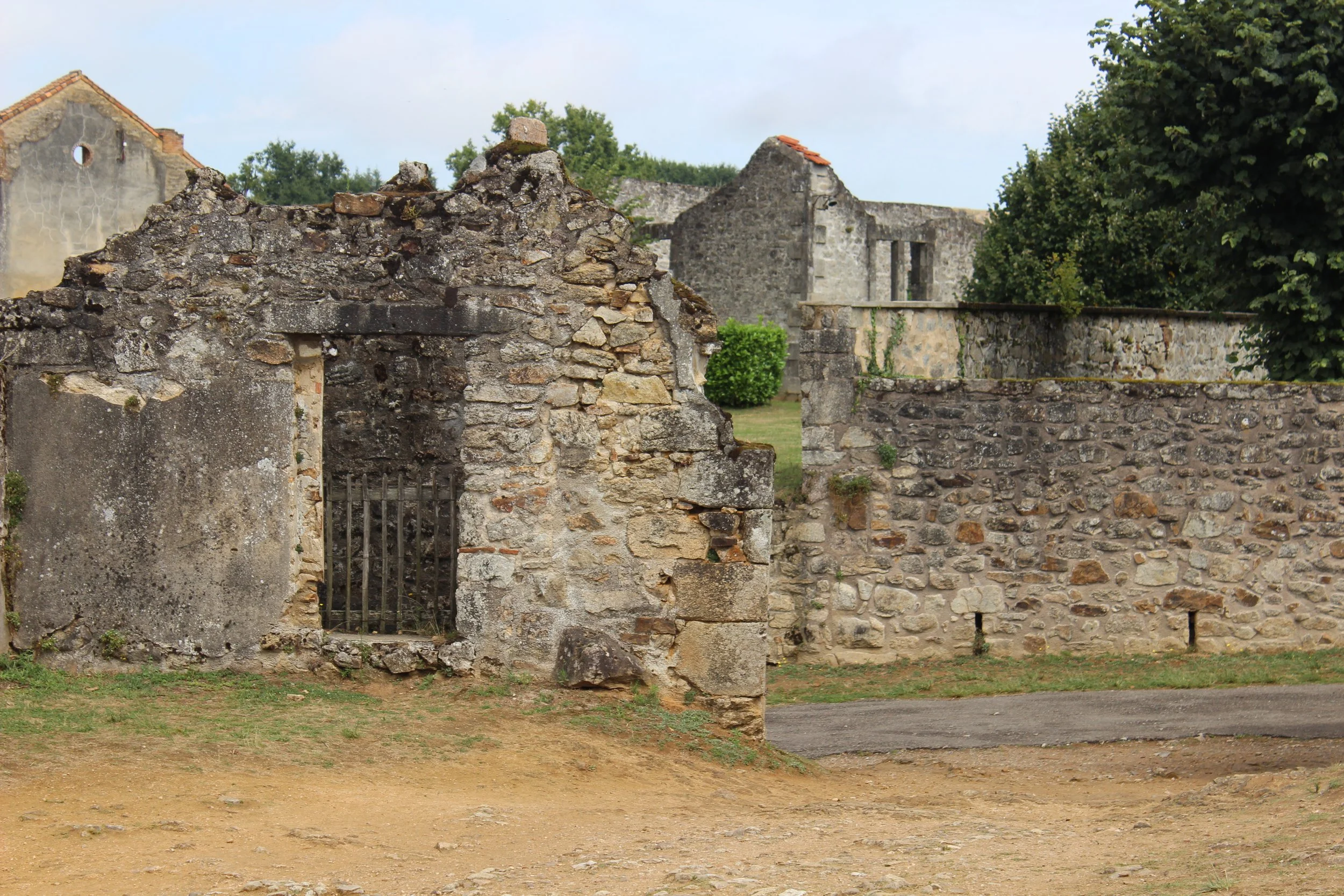 Verfallene Steinmauer mit Ritzen und einem kleinen, verschlossenen Gitter im Vordergrund, im Hintergrund alte Ruinen und Bäume.