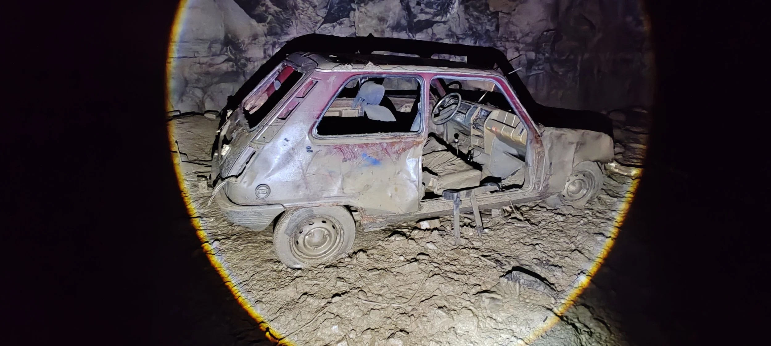Ein verrosteter, beschädigter und verlassen aussehender kleiner schwarzer Pkw in einer Höhle, umgeben von Felsen, beleuchtet vom Schein einer Taschenlampe.