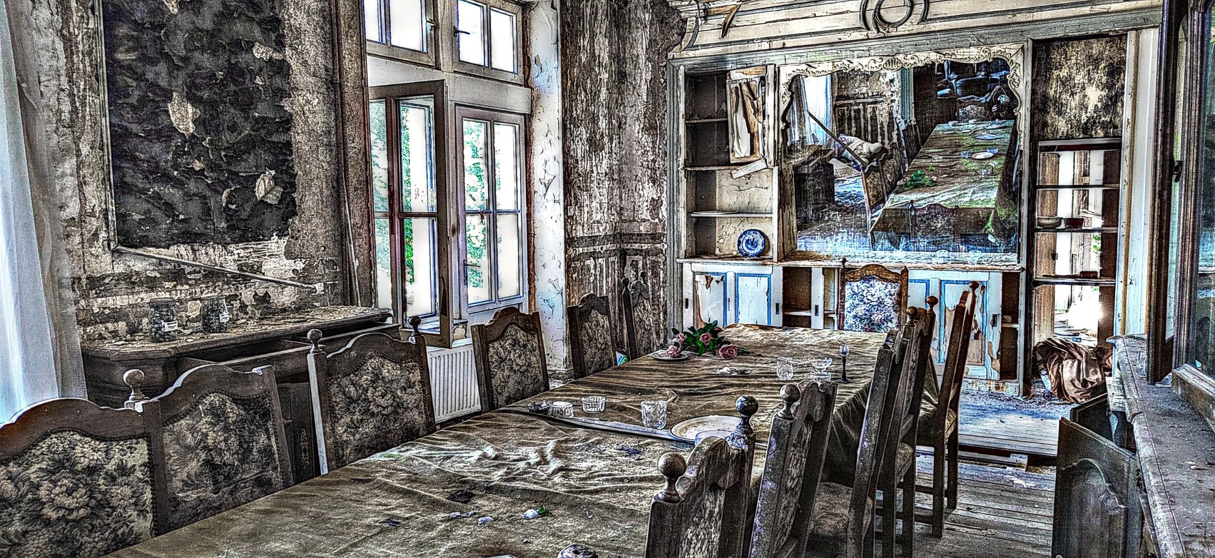Ein verlassener, alter Speisesaal mit einem großen Holztisch, umgeben von gepolsterten Stühlen, auf dem Tisch liegen Gläser, Dekorationen und Kerzen, die Wände sind beschädigt und die Fenster lassen viel Licht herein; im Hintergrund eine eingerichtet
