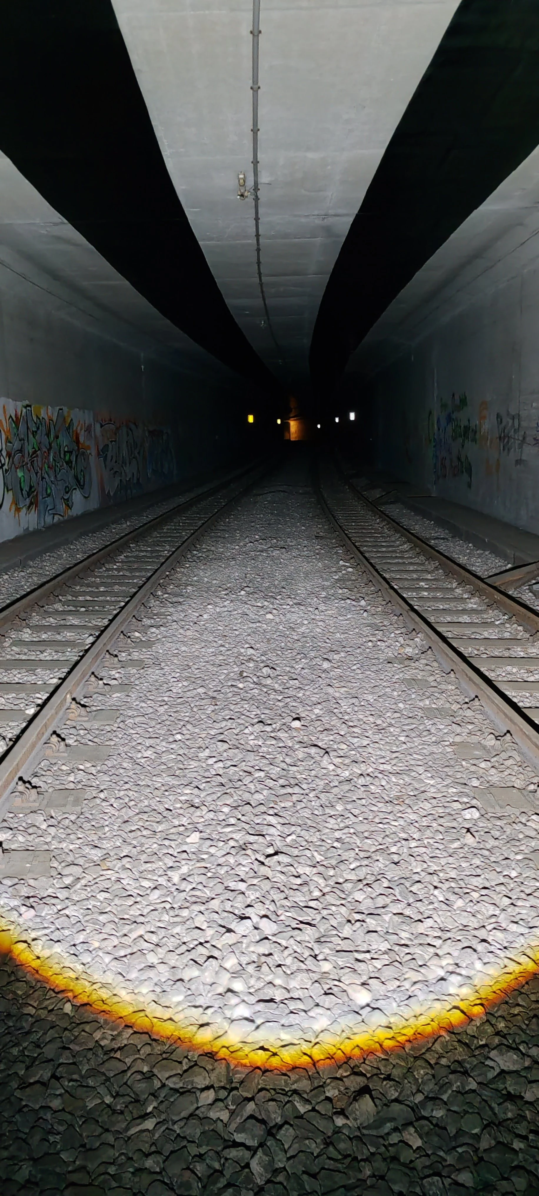Ein dunkler Eisenbahntunnel mit Graffiti an den Wänden, Gleise und Schotter im Vordergrund, am Ende des Tunnels ein Lichtpunkt.