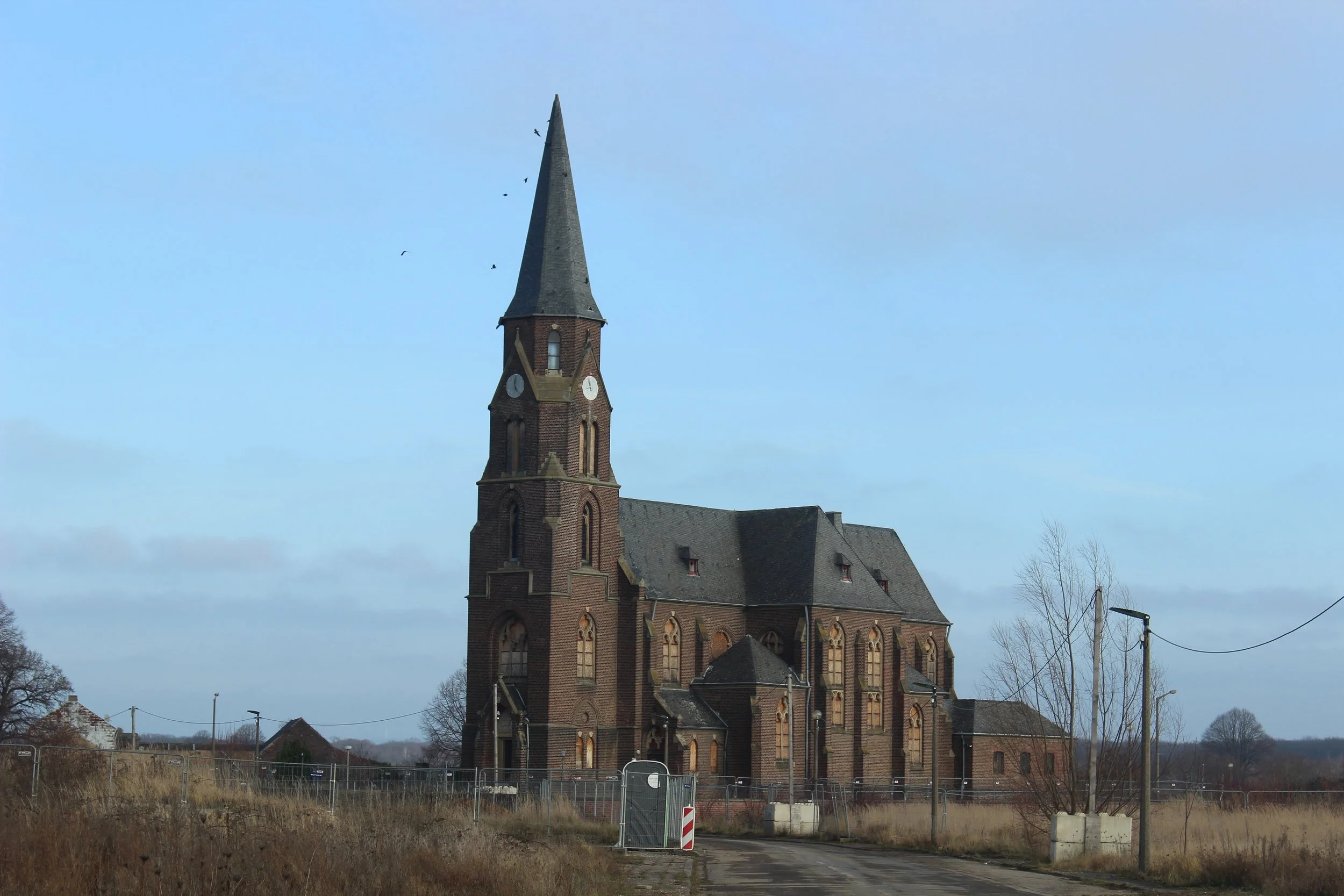 Eine alte, mittelgroße Kirche aus rotem Backstein mit hohem, spitzen Turm in einer ländlichen Umgebung, umgeben von Feld und vereinzelten Bäumen, mit einem Weg vor der Kirche und einem Bauzaun.