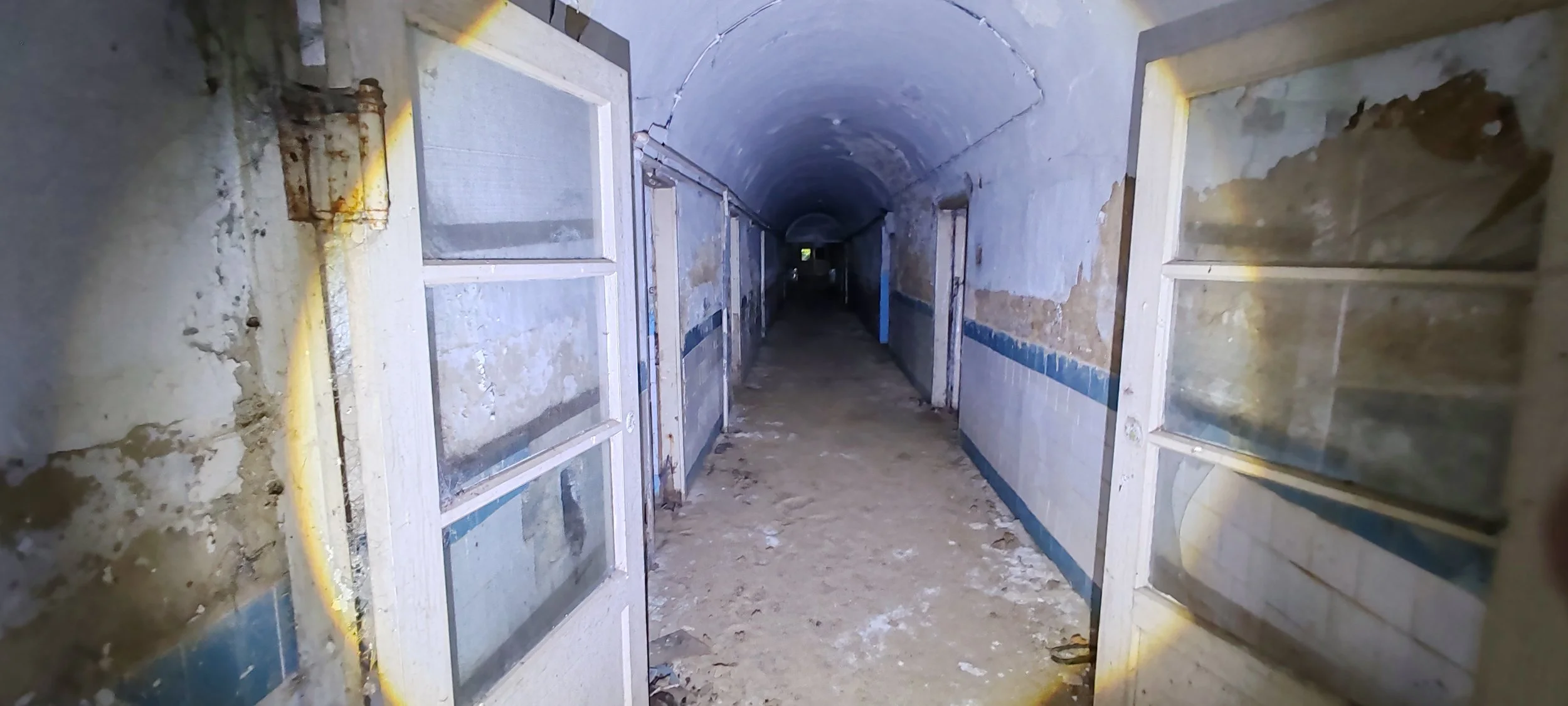 Ein dunkler, langen Flur in einem alten Gebäude, mit abgenutzten Wänden und veralteten Türen, die offenstehen.