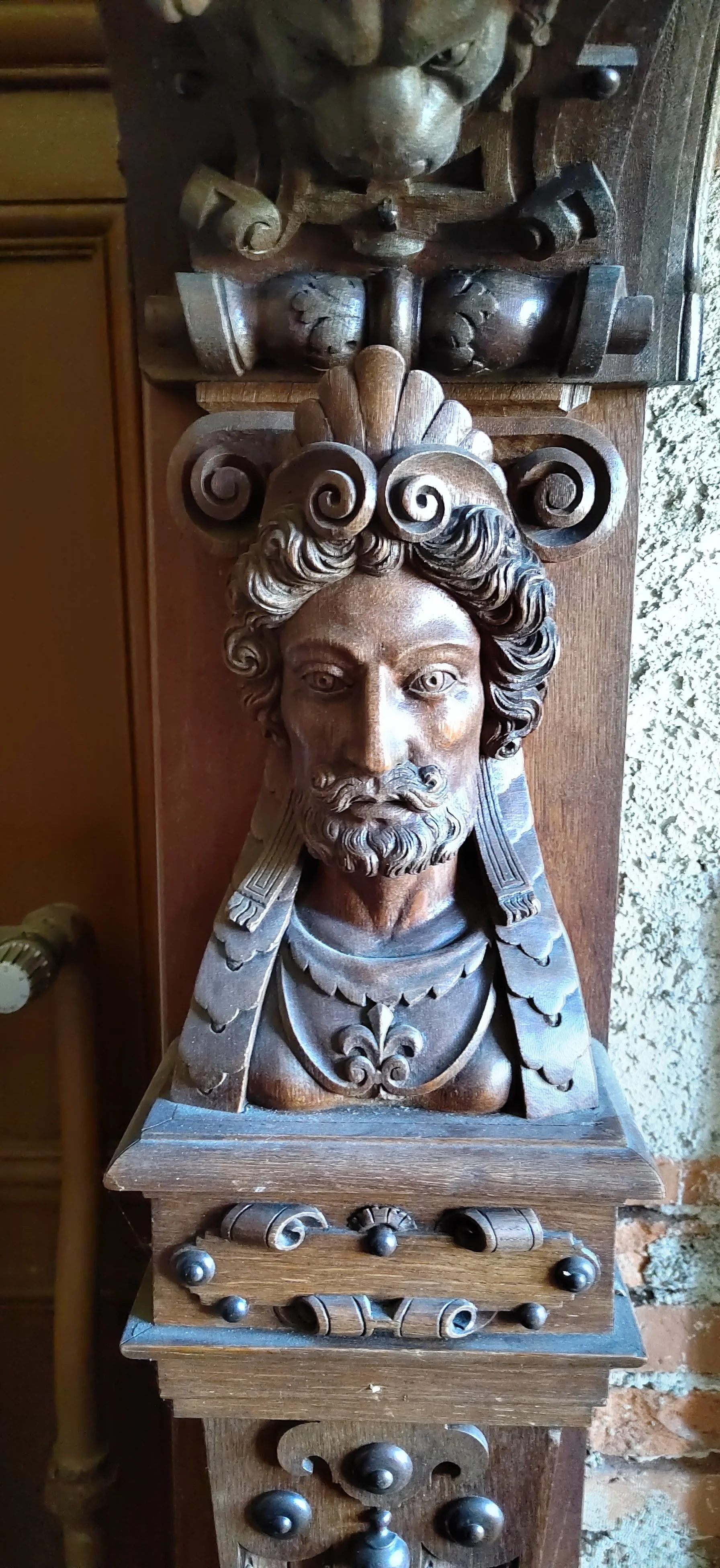 Holzskulptur eines historischen Mannes mit welligem Haar und Bart, verziert mit Ornamenten und auf einem kunstvoll geschnitzten Holzsockel.