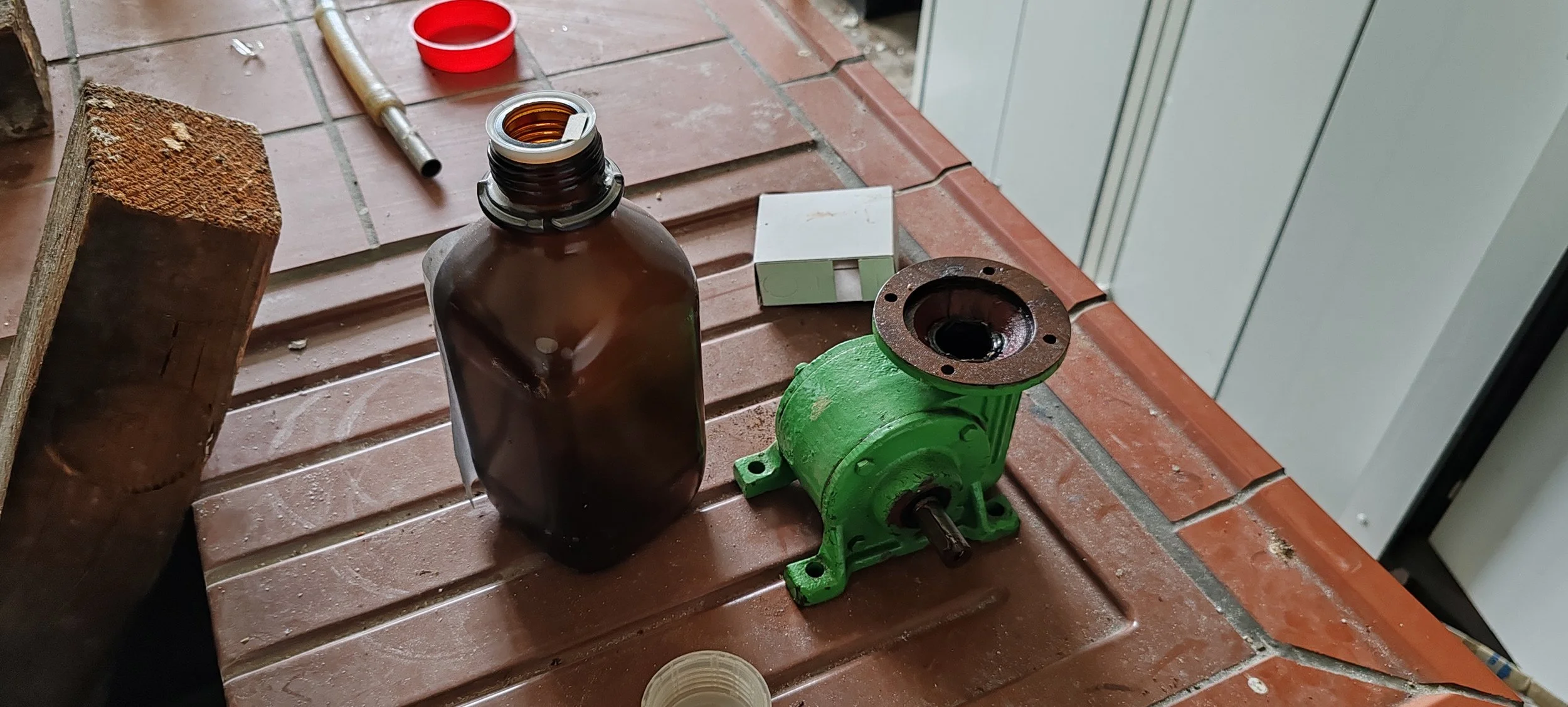 Baugruppe von Maschinen- oder Pumpenkomponenten, eine mit grün lackiertem Gehäuse, ein Fläschchen mit Öl, ein roter Deckel, und Werkzeuge auf einer roten Fliesenarbeitsfläche.