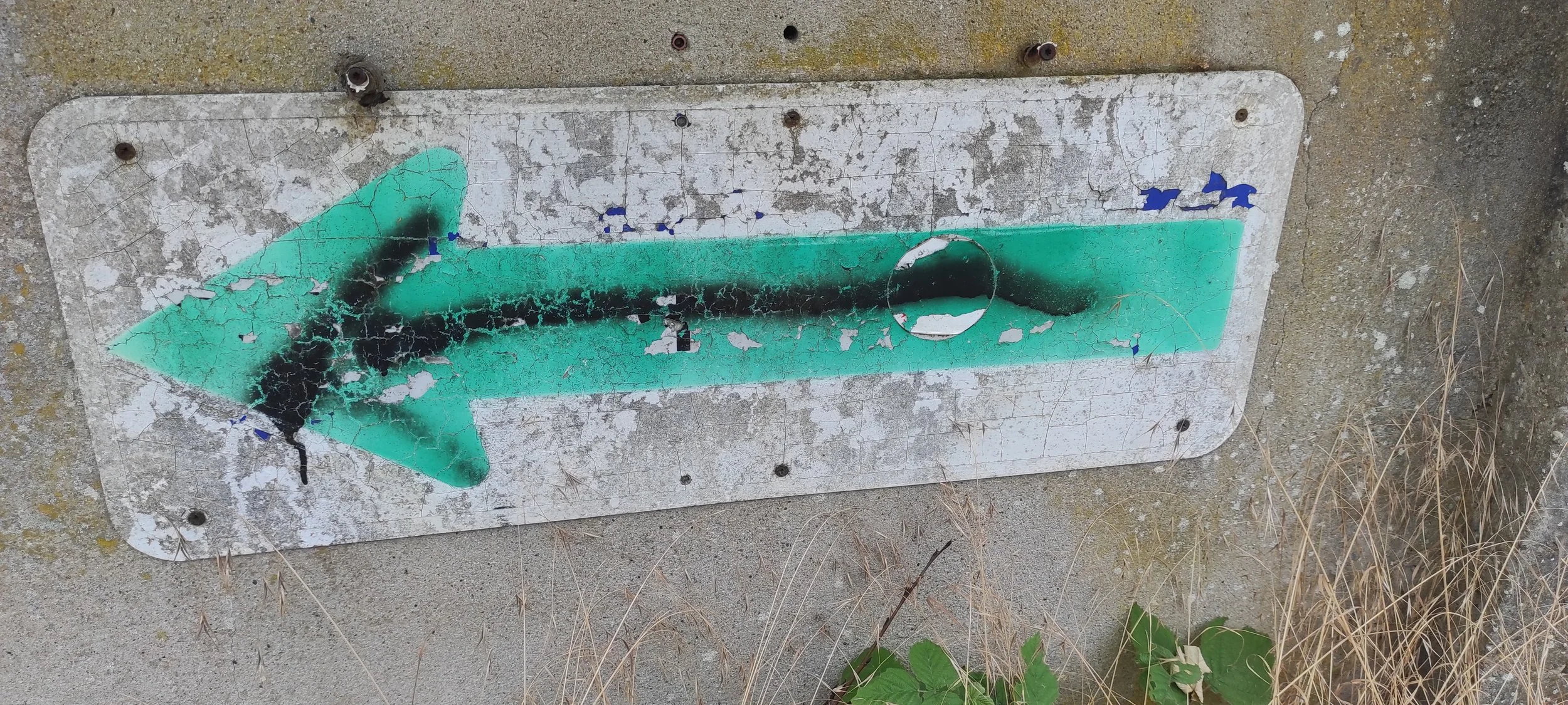 Verrostetes, beschädigtes Verkehrsschild mit grünem Pfeil, das nach links zeigt, auf einer staubigen Wand, umgeben von trockenen Gräsern.