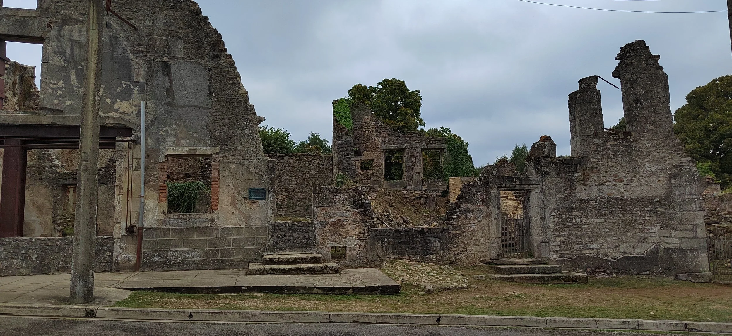 Ruinen eines alten Gebäudes, teilweise von Pflanzen überwuchert, mit einigen Stufen vor einer Straße, bei bewölktem Himmel.