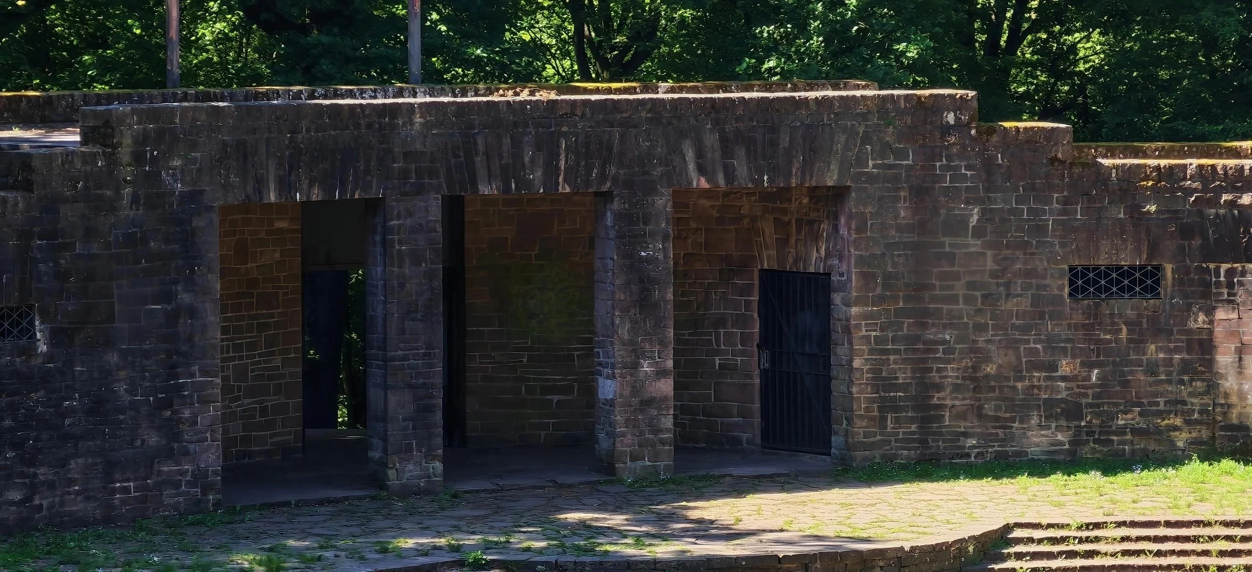 Eine alte Steinmauer mit Tor und kleinen Fenstern, umgeben von grünen Bäumen, im Freien. Der Boden ist gepflastert.
