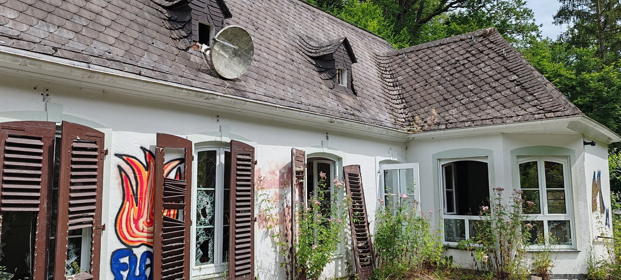 Ein altes, weißes Haus mit braunen Jalousien, Graffiti an der Wand, kaputte Fenster, und einem Schrägdach mit Dachziegeln. Im Vordergrund sind Blumen und Pflanzen.