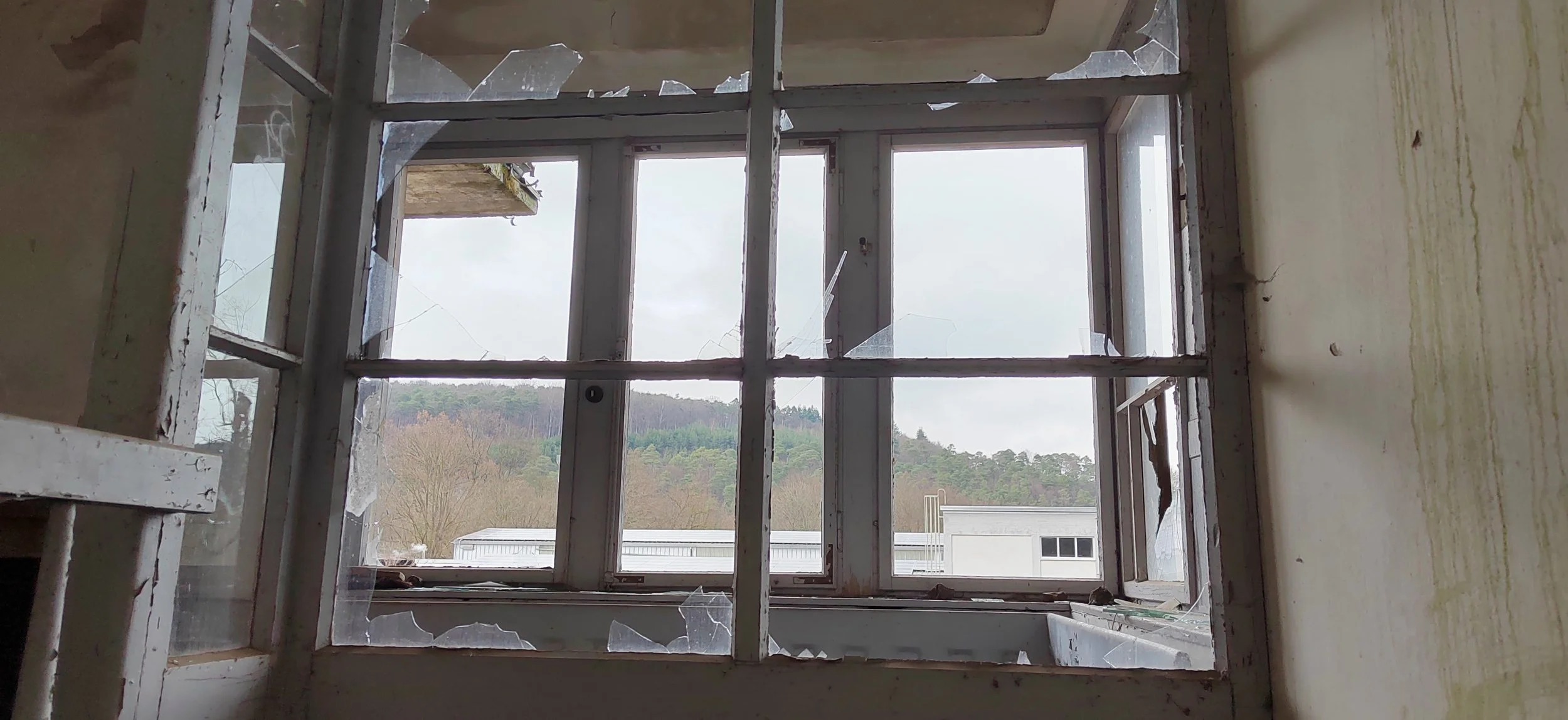 Ein zerbrochener Fensterrahmen in einem Raum, durch den man eine Landschaft mit Bäumen und Hügeln im Hintergrund sieht.
