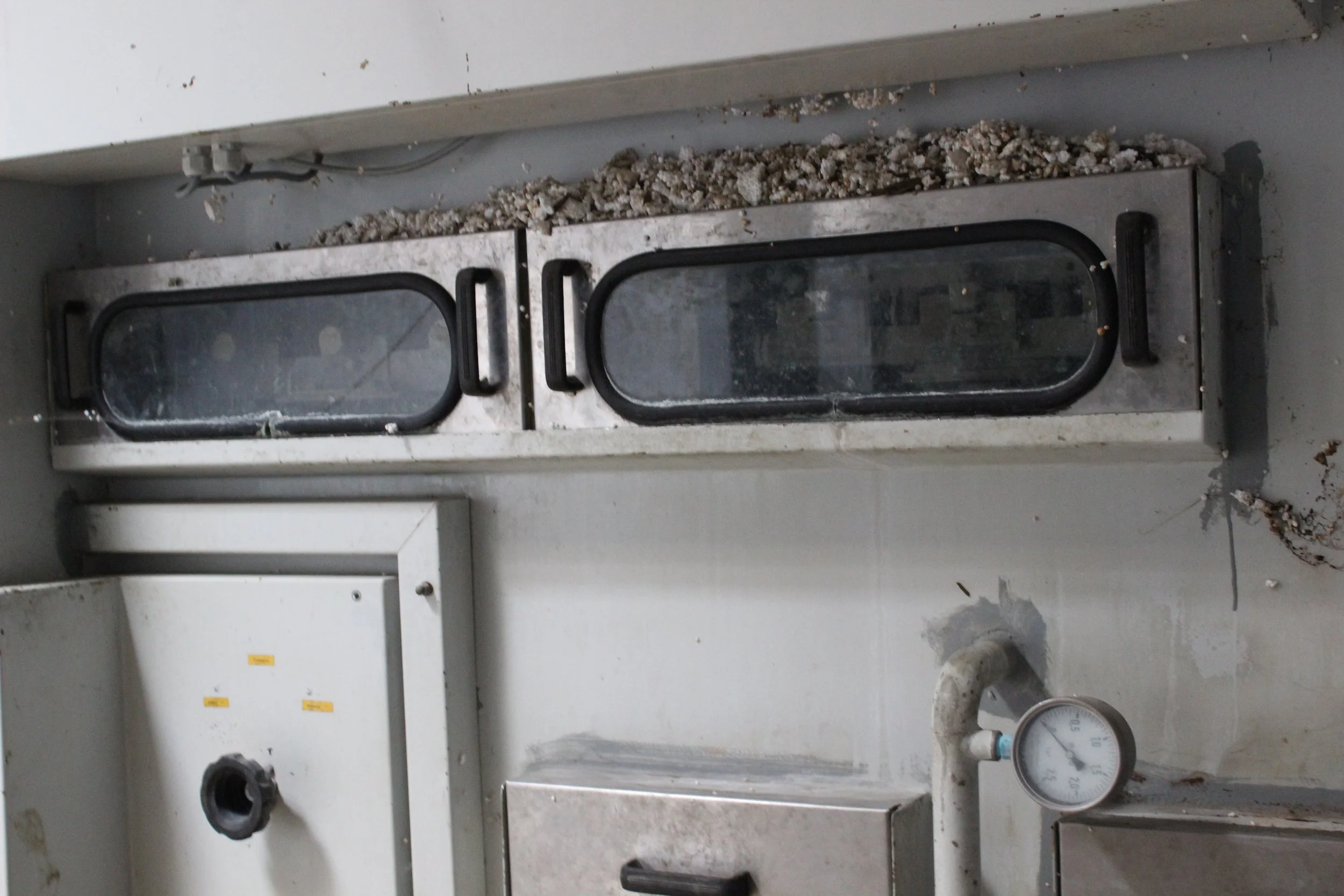 Blick auf eine alte medizinische oder laborausstattung, mit einem Thermometer im Vordergrund und einer Klappe am Boden, die mit Schotter bedeckt ist, alles wirkt veraltet und benutzt.