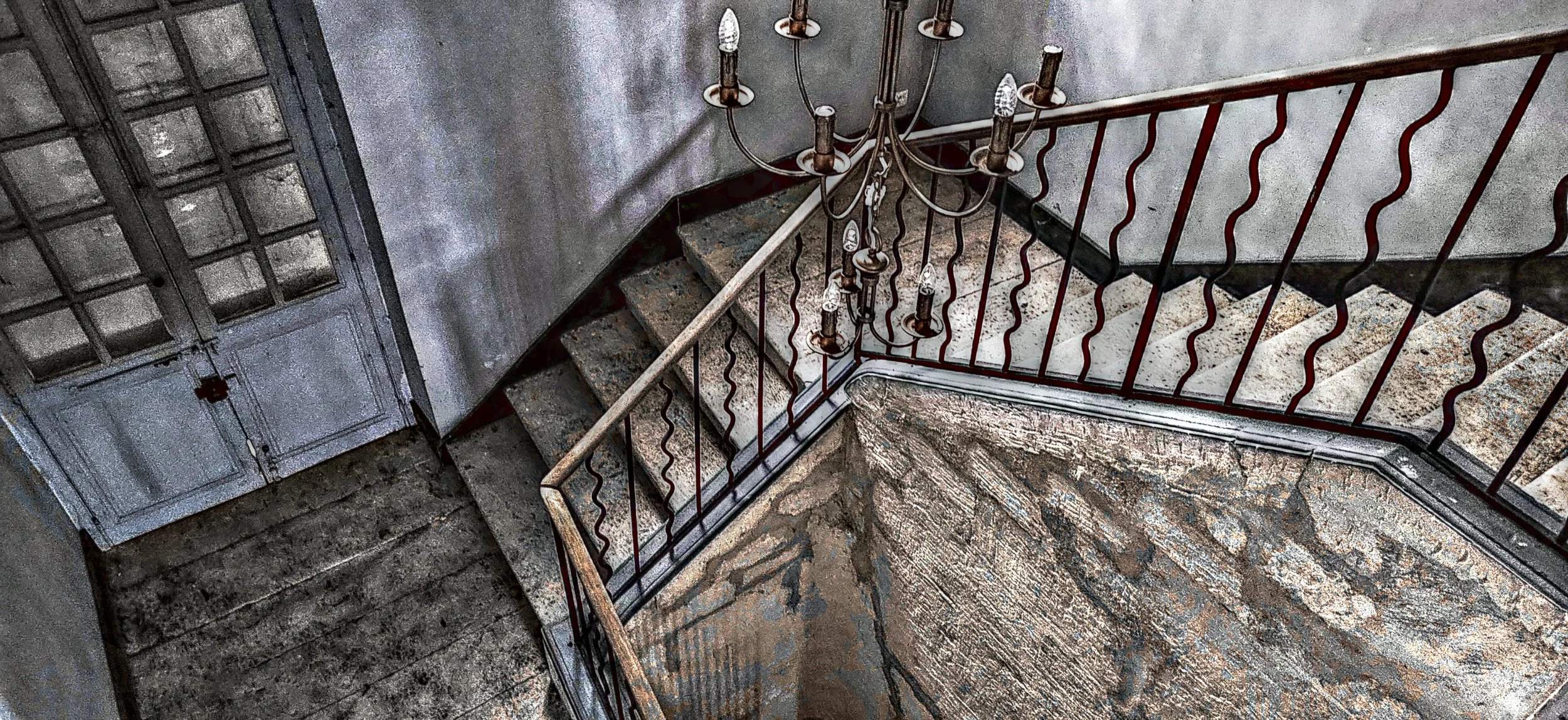 Ein Blick nach oben auf eine antike, schlichte Treppe mit Steinstufen, einer rustikalen Wand aus Backstein, einer alten Metallkugel-Kronleuchte und eine alte Tür.