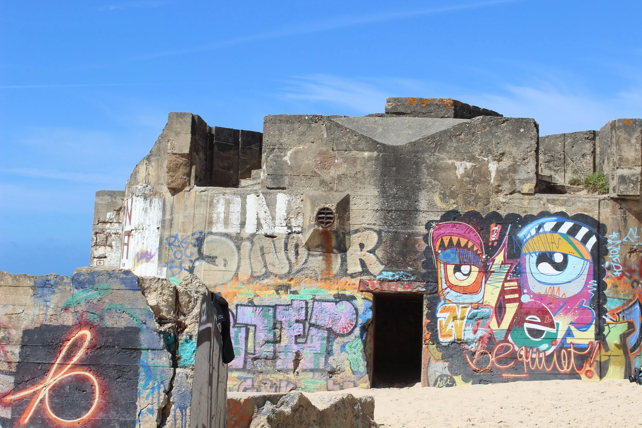 Graffiti an einer alten Betonmauer unter blauem Himmel, mit einer offenen Tür und bunten, stilisierten Augen in der Street Art