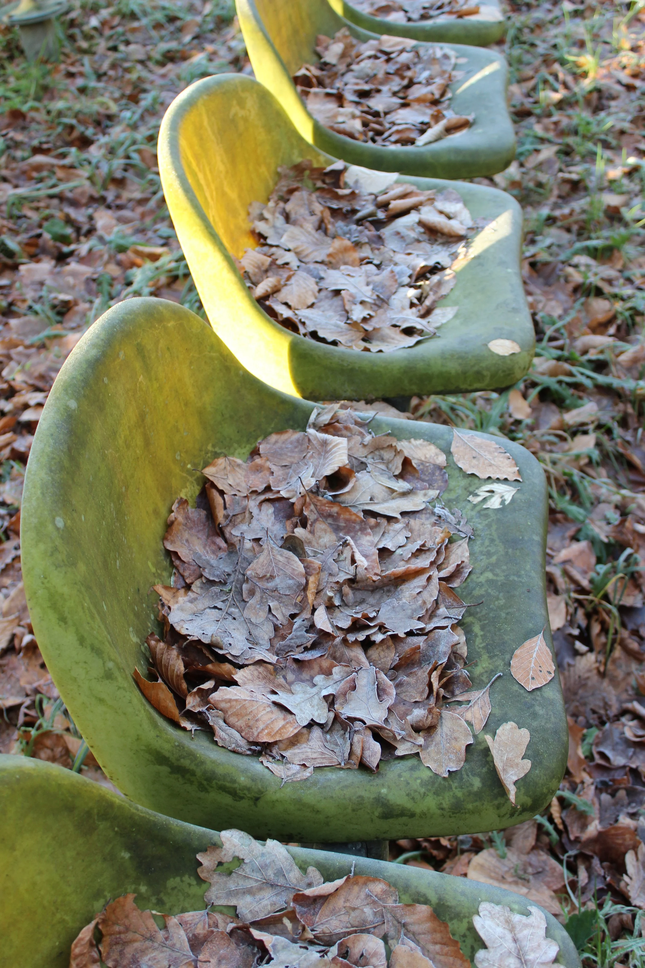 Mehrere grüne Massagesessel im Park, mit trockenen Blättern bedeckt.