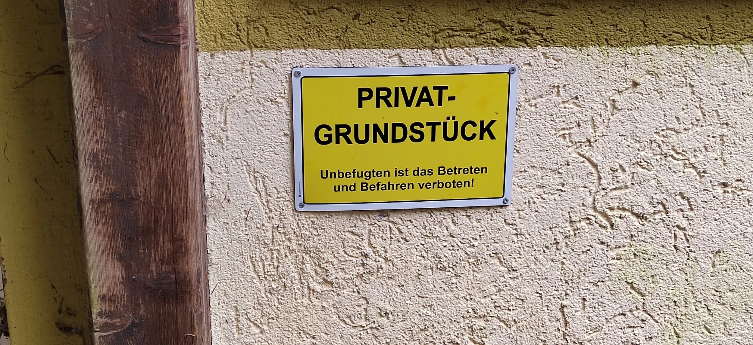 Ein Schild an einer Hauswand mit gelbem Hintergrund, schwarzer Umrandung und schwarzer Schrift, das den Zugang zu einem Privatgrundstück verbietet.