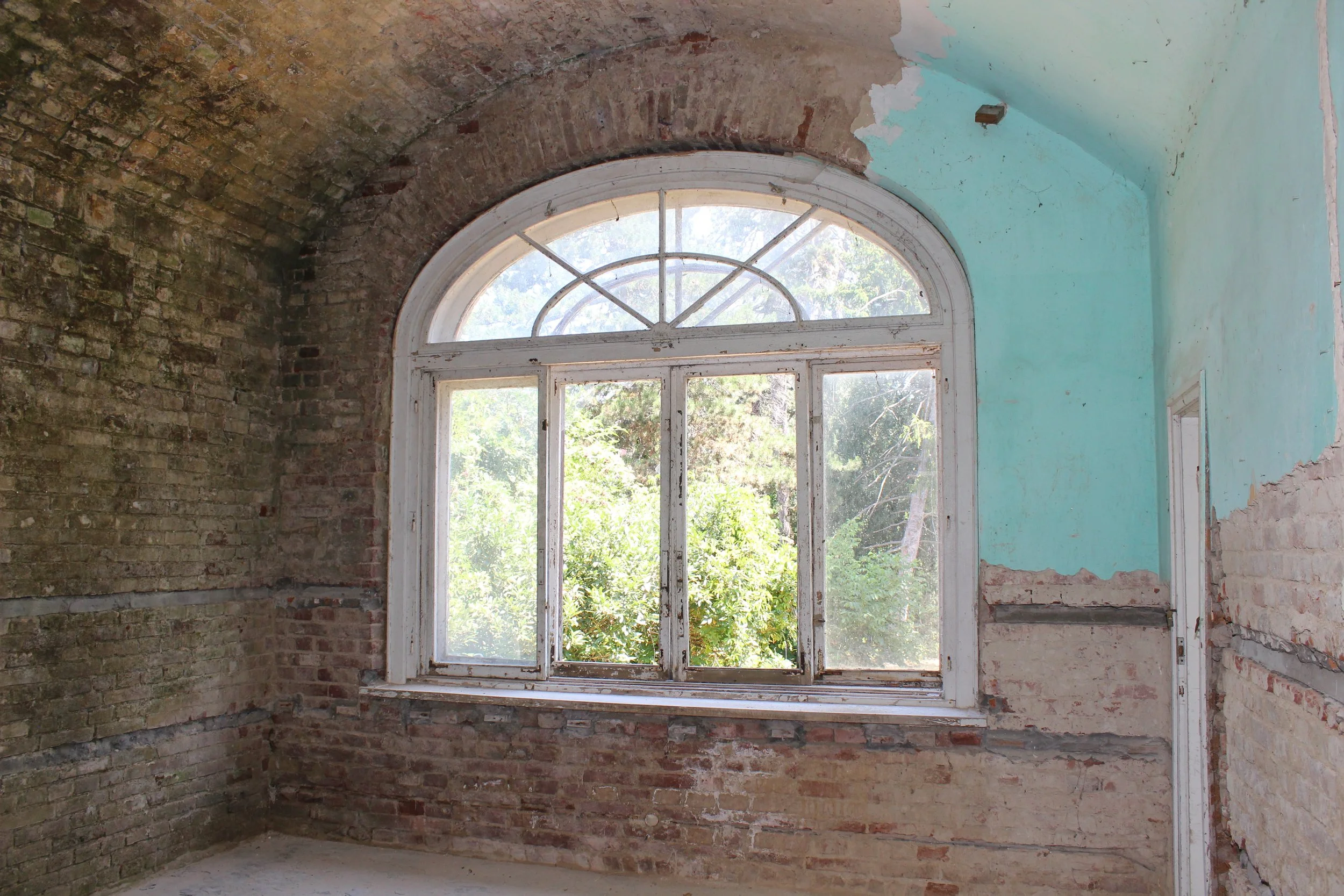 Ein alter, verlassener Raum mit großen, weißen Fensterrahmen und Blick nach draußen auf Bäume. Wände sind teilweise verputzt und teilweise sichtbar mit alterte Backsteinstruktur. Das Dach ist gewölbt.