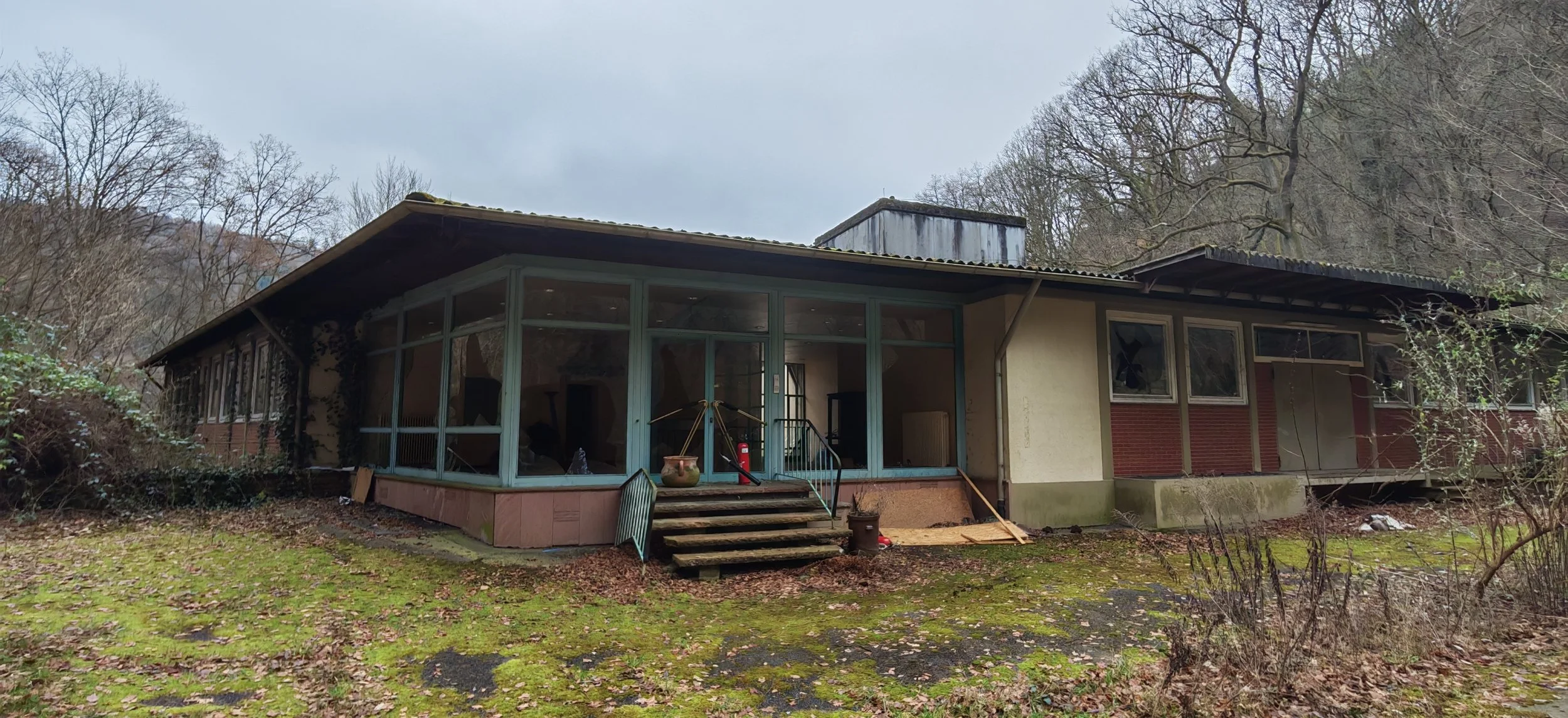 Verlassenes Gebäude im Wald mit verfallener Fassade, zerbrochenen Fenstern und moosbedecktem Boden, umgeben von Bäumen.