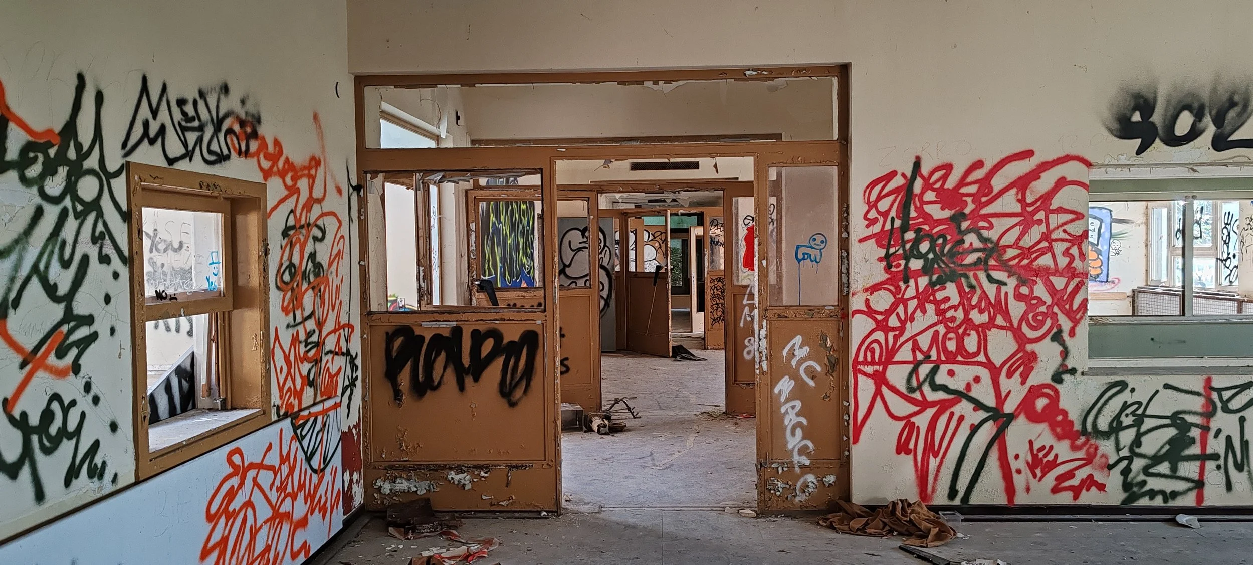 Innenansicht eines verlassenen, verfallenen Gebäudes mit Graffiti an den Wänden, zerbrochenen Fenstern und zerfetztem Bodenbelag.