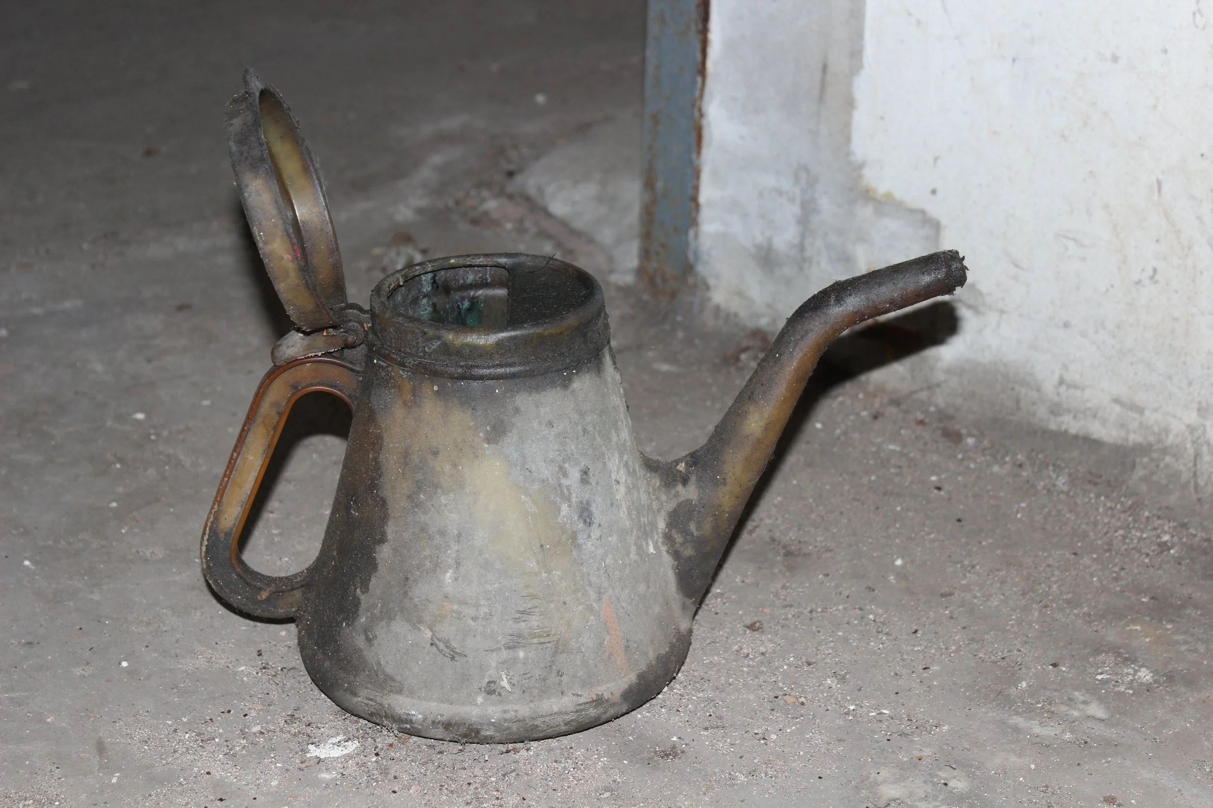 Ein verrosteter, alter Wasserkrug mit geöffnetem Deckel liegt auf einer staubigen, verschmutzten Bodenfläche in einer unordentlichen Umgebung neben einer Wand.