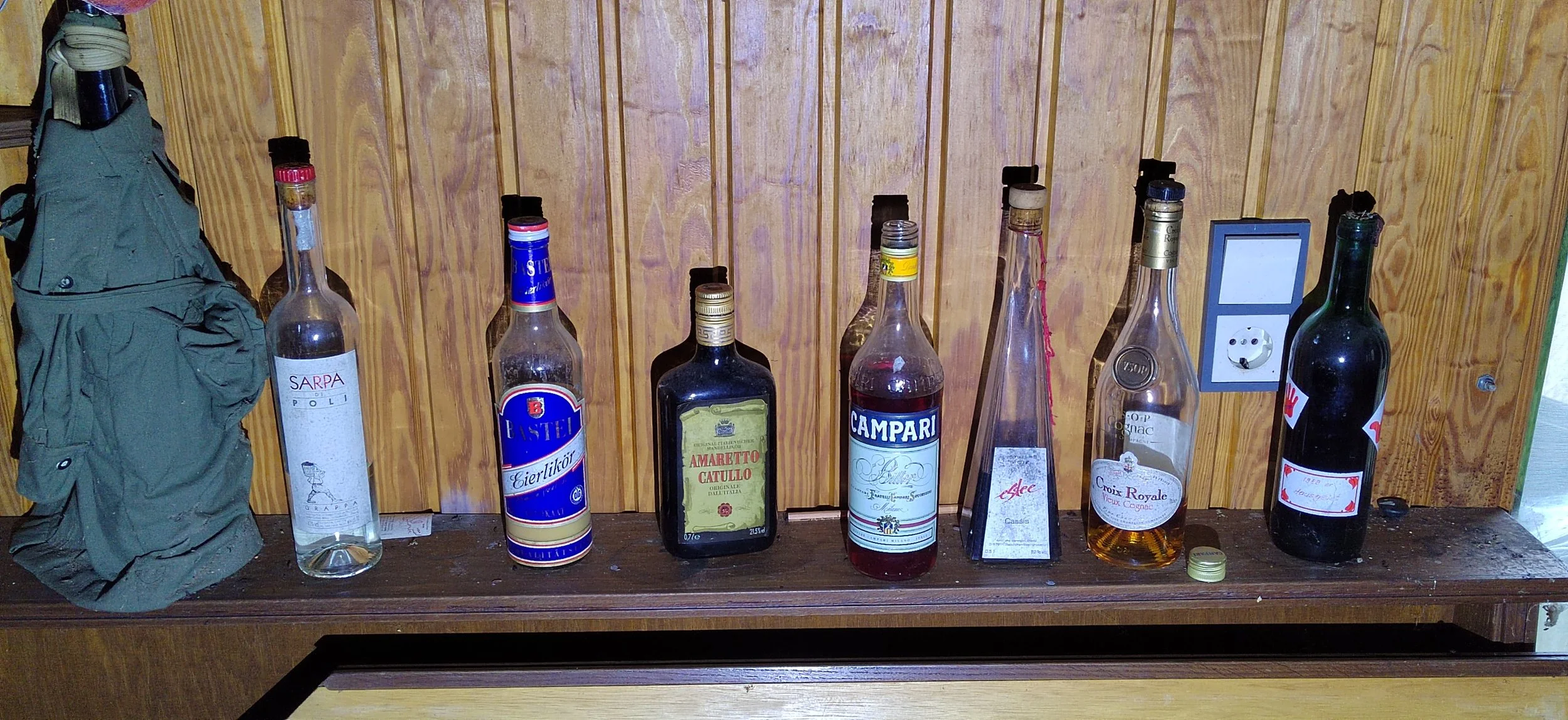 Verschiedene leere Alkoholflaschen auf einem Holzregal, einschließlich Wodka, Amaretto, Campari, Cognac und Wein, vor einer holzvertäfelten Wand.