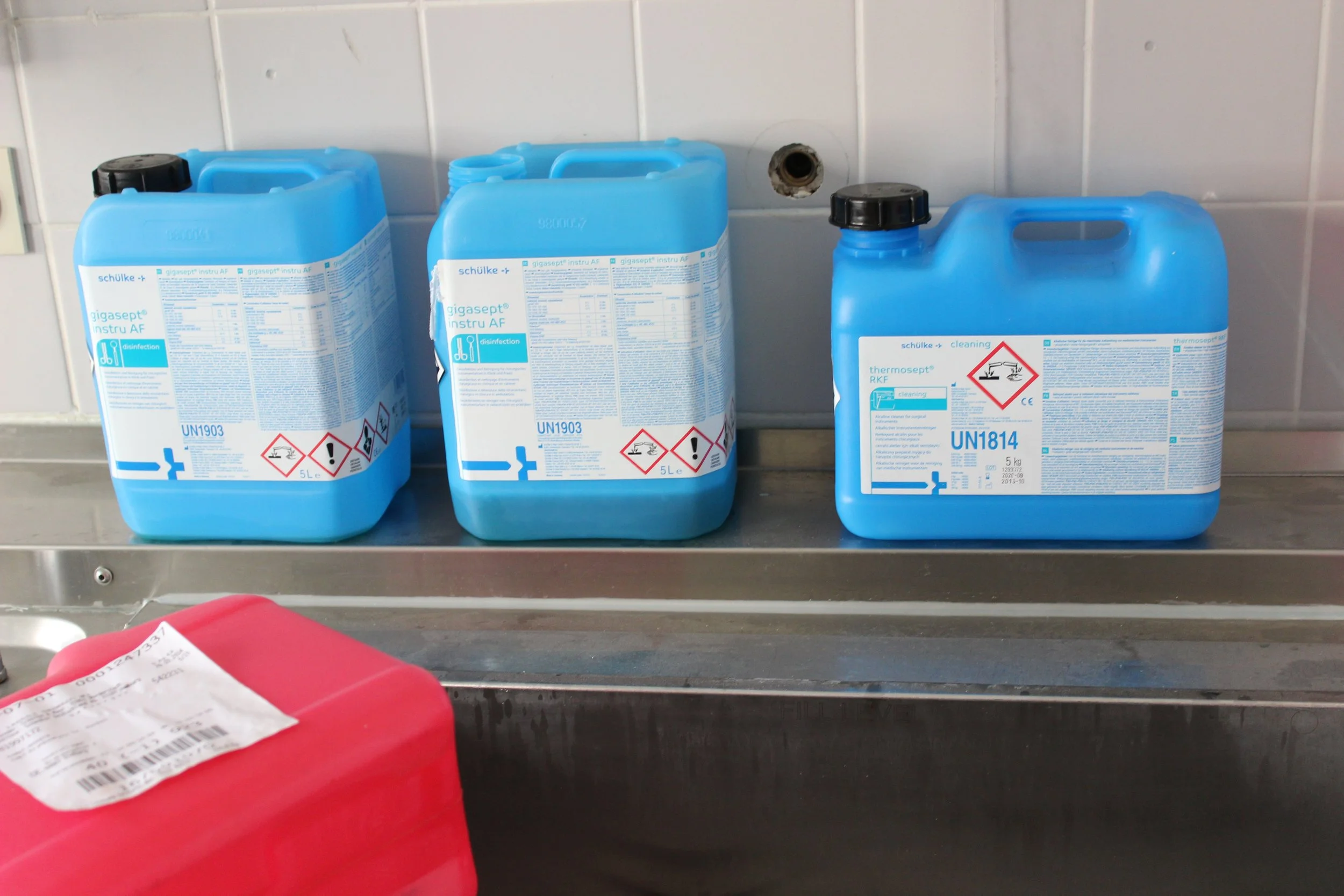 Drei blau Plastikbehälter für Reinigungsmittel stehen auf einer Metallablage, mit einem roten Behälter im Vordergrund. Die Behälter haben Warnsymbole und Produktinformationen.