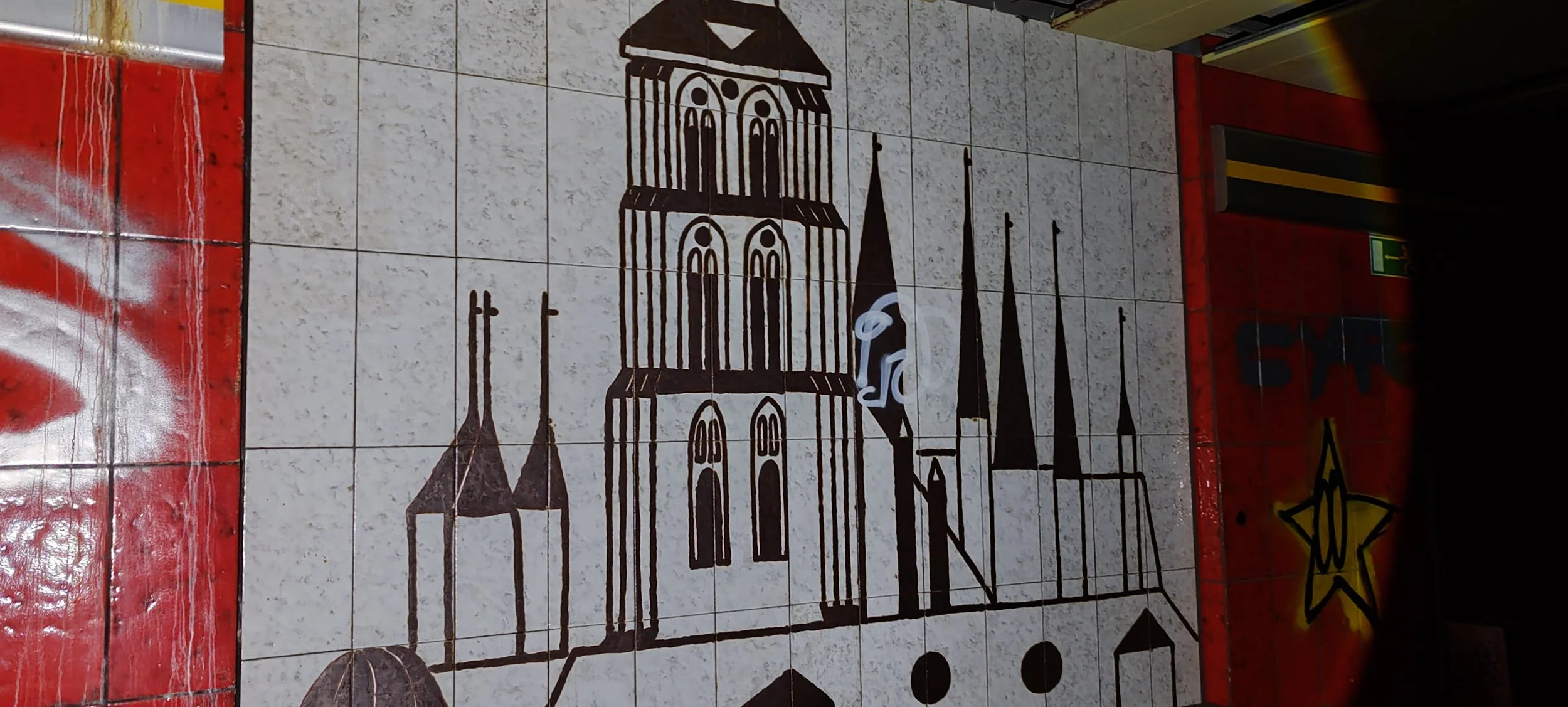 Wandfliese mit schwarz-weißem Bild eines Kirchengebäudes mit Türmen und Spitztürmen, rote Fliesen um die Fliese, ein gelber Stern auf schwarzem Hintergrund, ein grüner Notausgangsschalter auf der Wand.