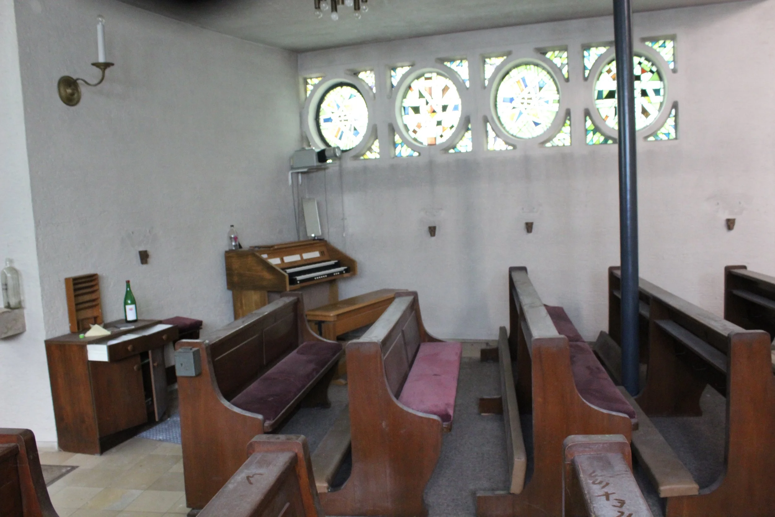 Innenraum einer kleinen Kirche oder Kapelle mit Holzbänken, bunten Glasfenstern und einem Orgelpult.