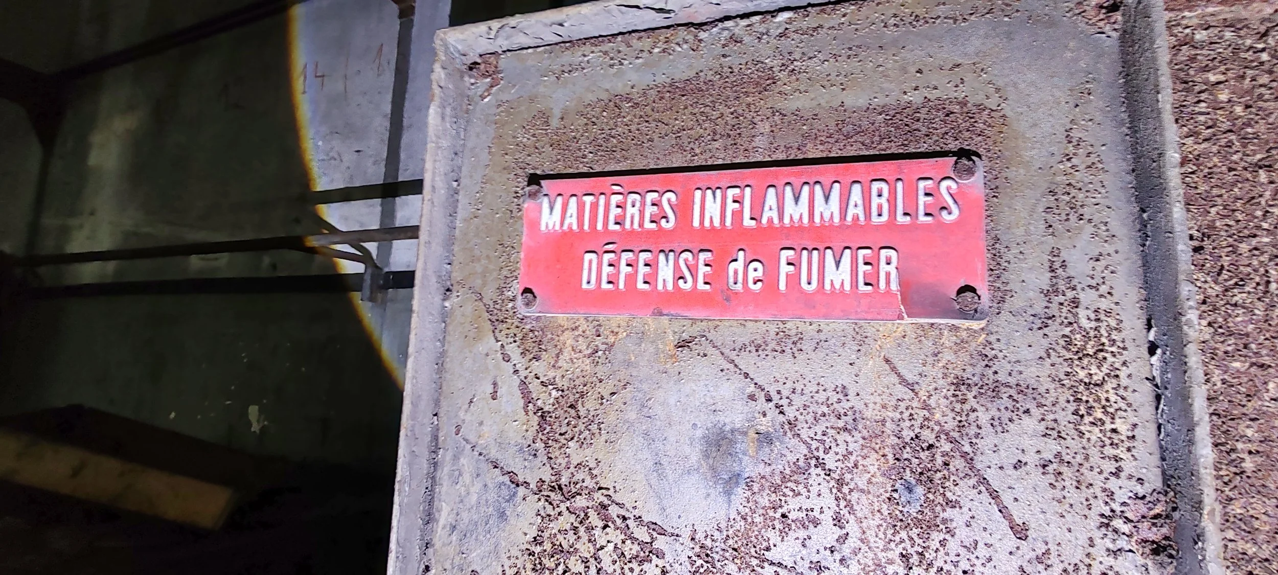 Rostfreie Metallplatte mit roten Hintergrund und weißen Buchstaben, die auf einem Betonfundament angebracht ist. Der Text lautet: "MATIERES INFLAMMABLES DÉFENSE de FUMER".