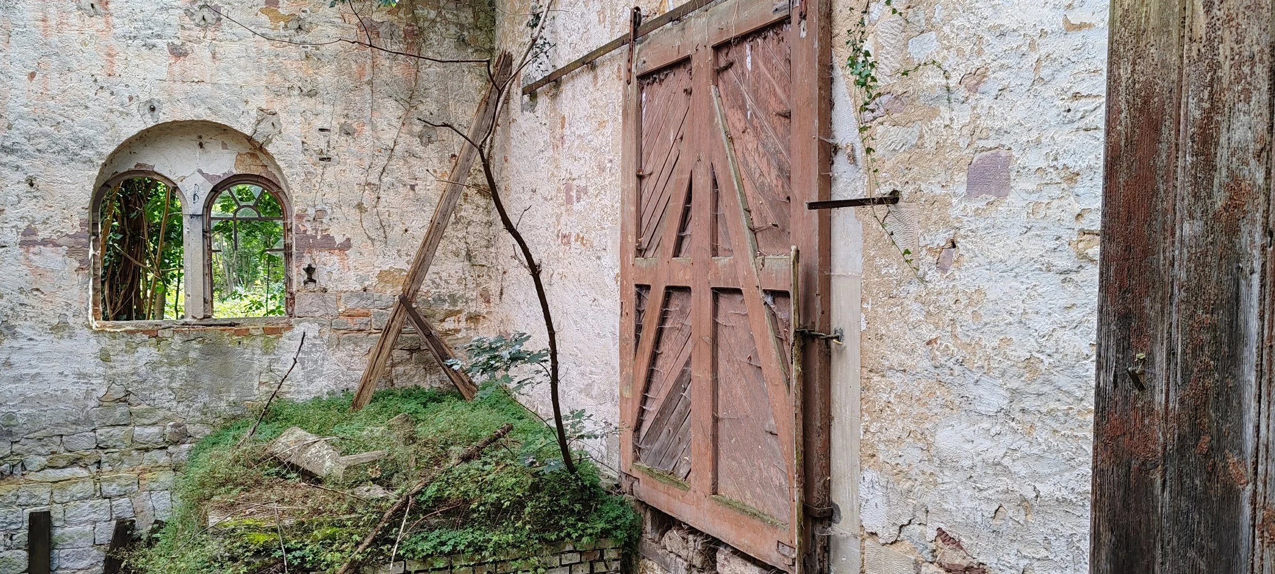 Ein verfallenes Haus mit einer renovierten Fensteröffnung und einem großen, alten Holztür, die teilweise offen steht. Die Wand ist alt und abgenutzt, mit sichtbaren Steinen und Efeu, das wächst. Draußen sind Bäume und Pflanzen sichtbar.