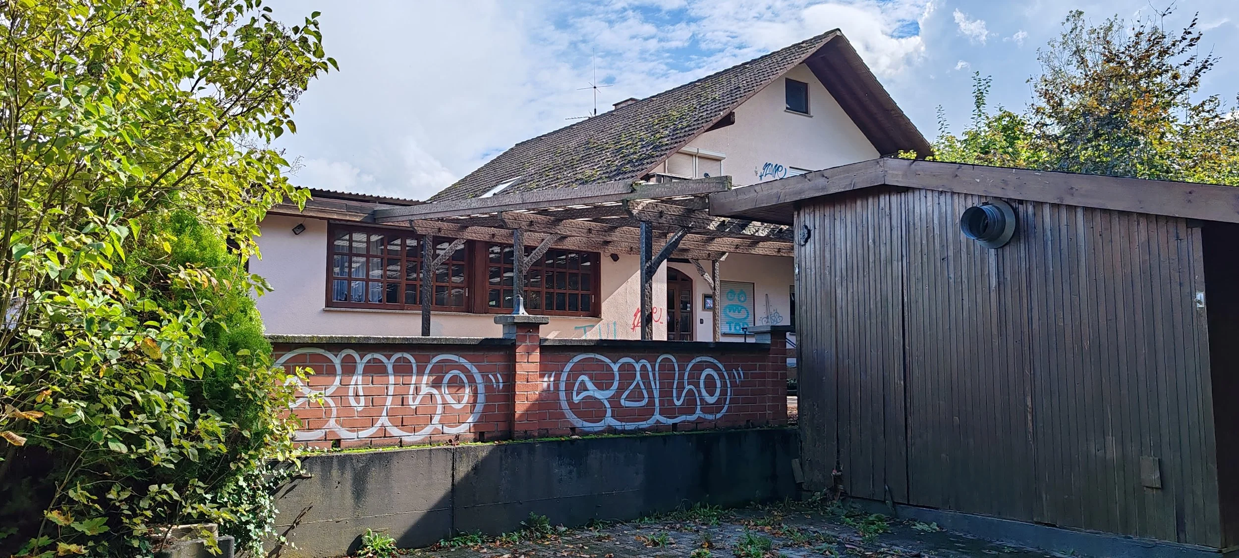 Ein Haus mit grauem Dach, braunem Holzapfel und Graffiti an der Wand, umgeben von Bäumen und einem blauen Himmel.