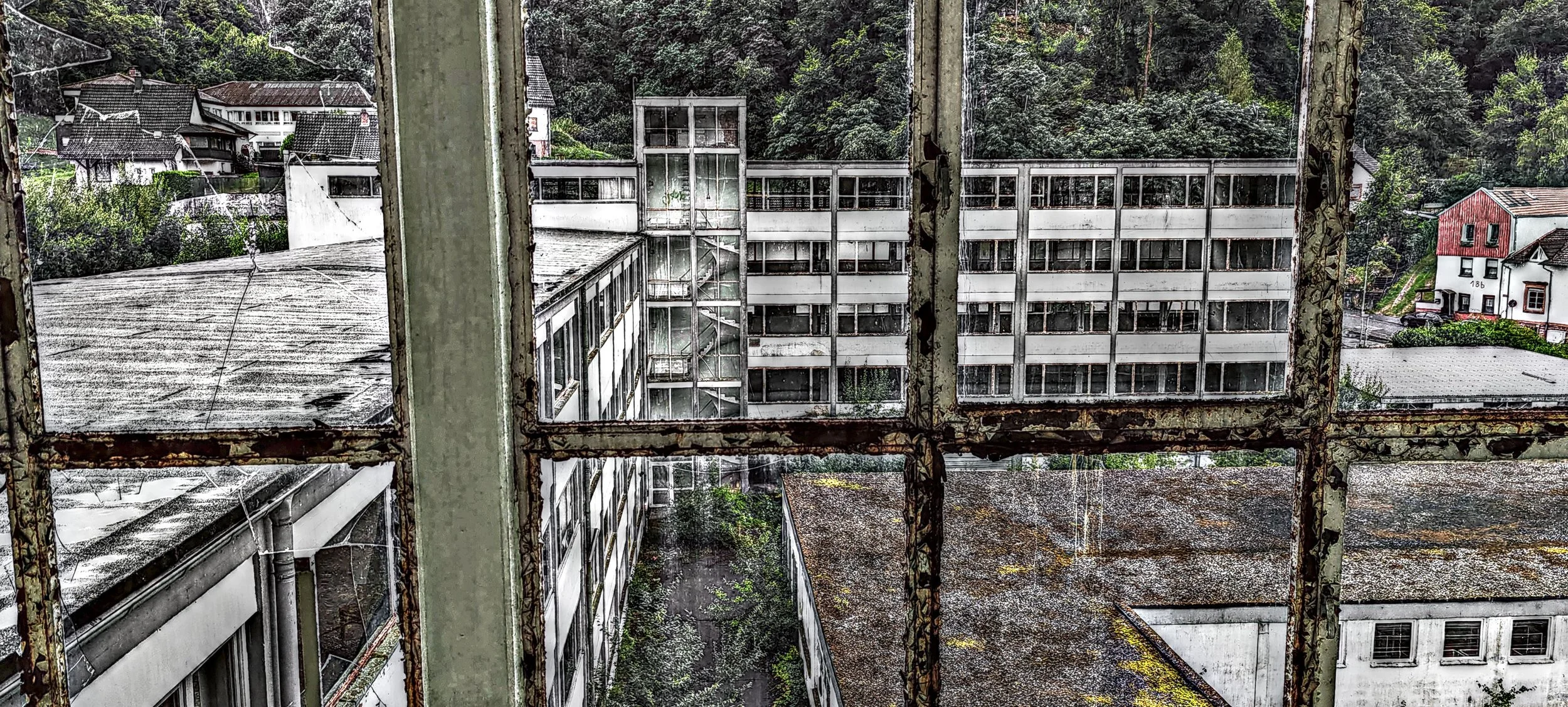 Blick durch verrostete Fenster auf eine verlassene, verfallene Gebäudefläche mit Gebäuden im Hintergrund, umgeben von grünen Hügeln und Bäumen.
