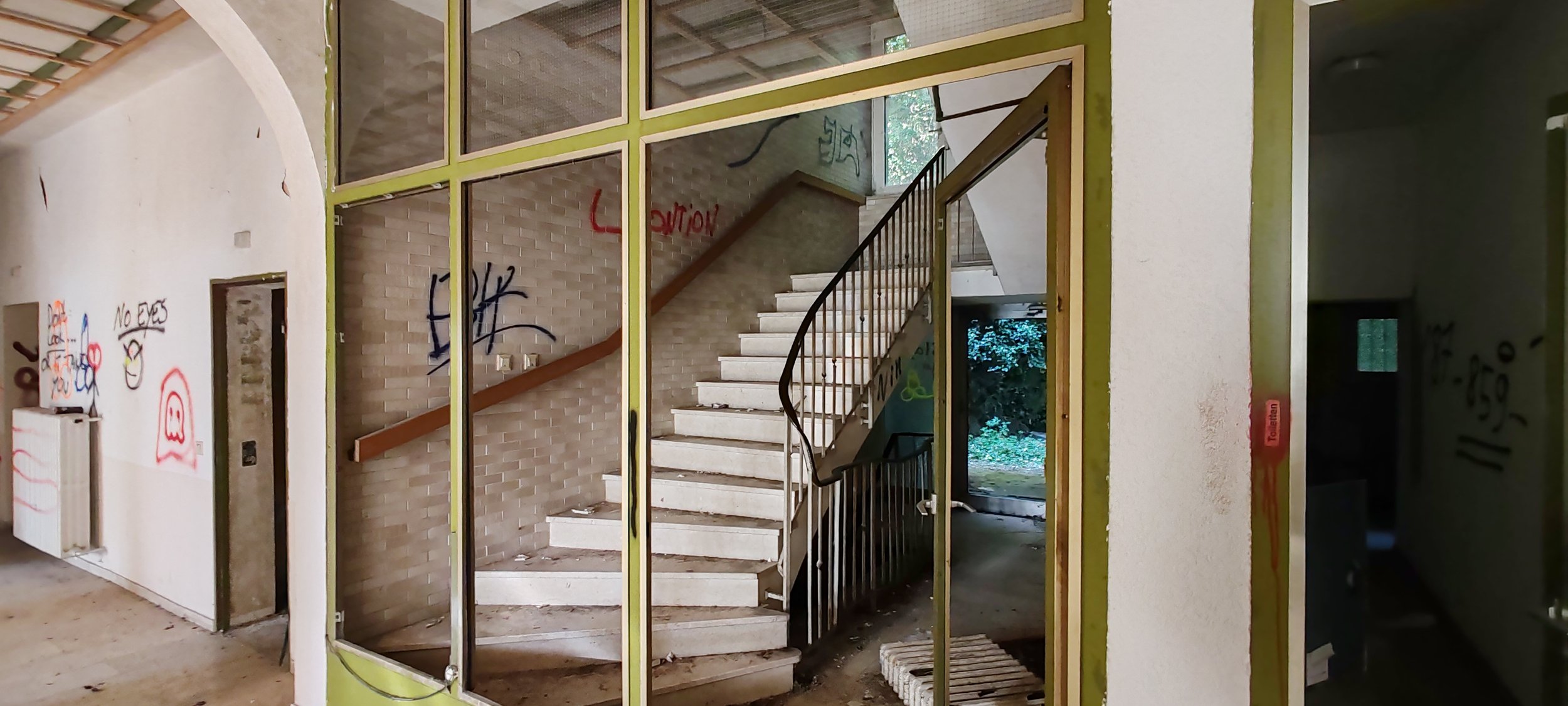 Innenansicht eines verlassenen Gebäudes mit einer Treppe, Graffiti an den Wänden und unordentlichen Boden. Glaswände trennen den Raum vom Flur.