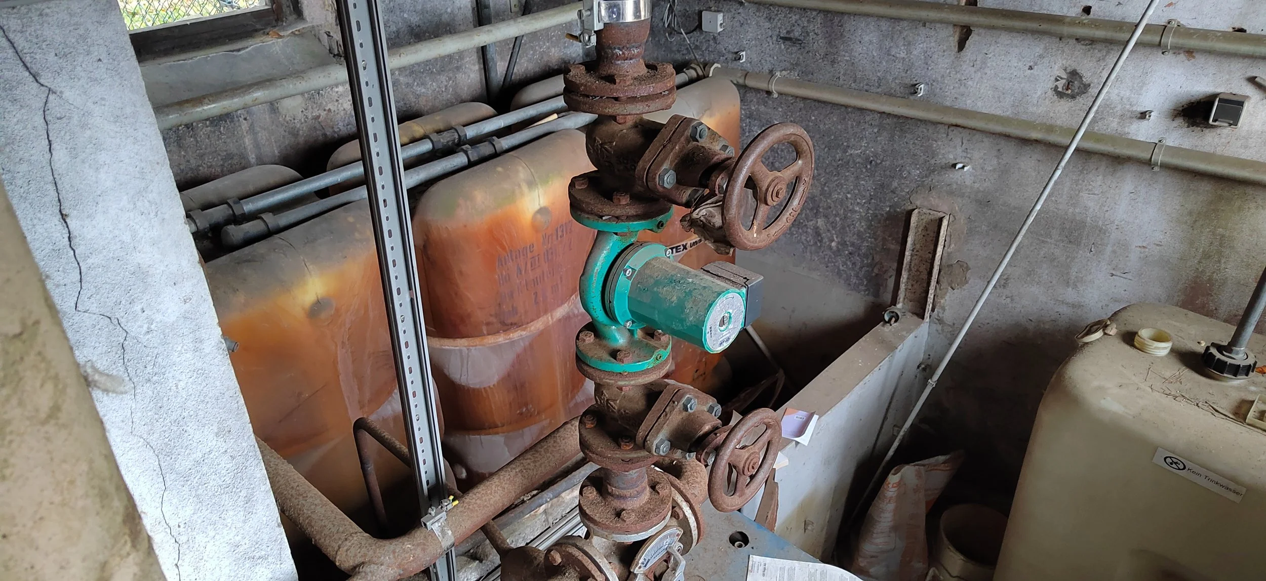 Rostige Wasserleitungen, ein Ventil mit Handrad und große Metallbehälter in einem unbelebten, alten Kellerraum.