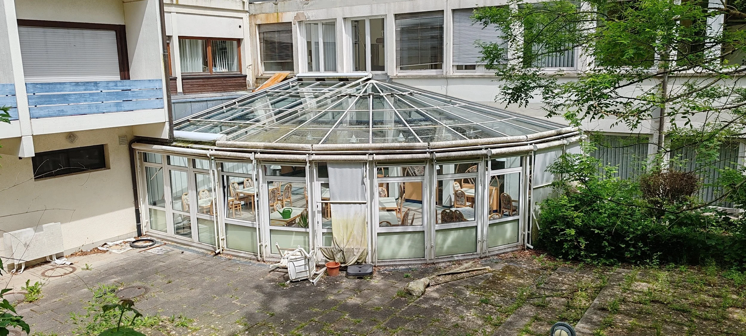 Ein alter Wintergarten aus Glas und Aluminium vor einem mehrstöckigen Wohngebäude. Im Inneren sind Tische und Stühle zu sehen, draußen befinden sich Pflanzen und Steine auf dem Boden.